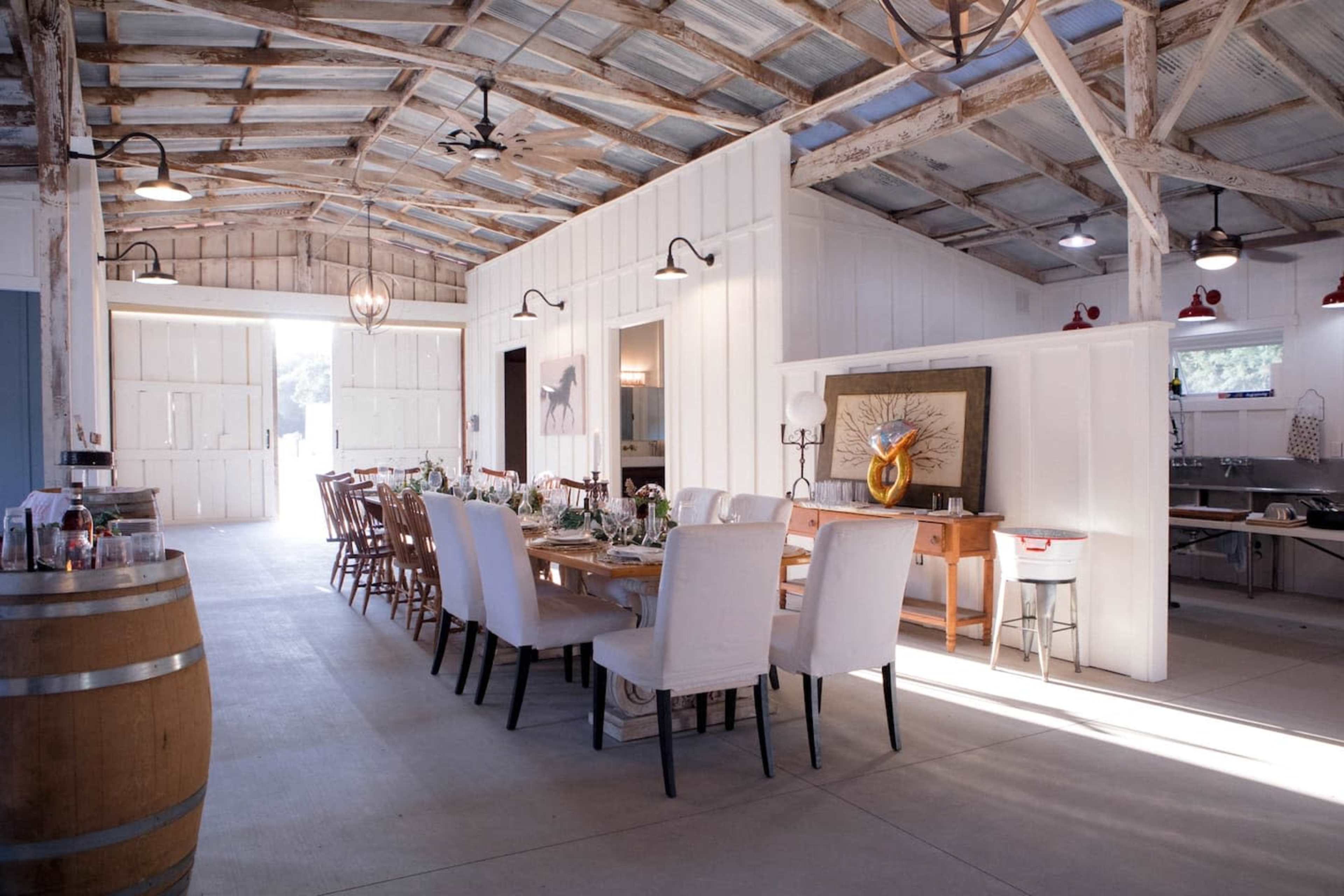 A spacious, rustic dining room with long wooden tables set for a meal, surrounded by white chairs, within a building featuring exposed beams and large double doors.