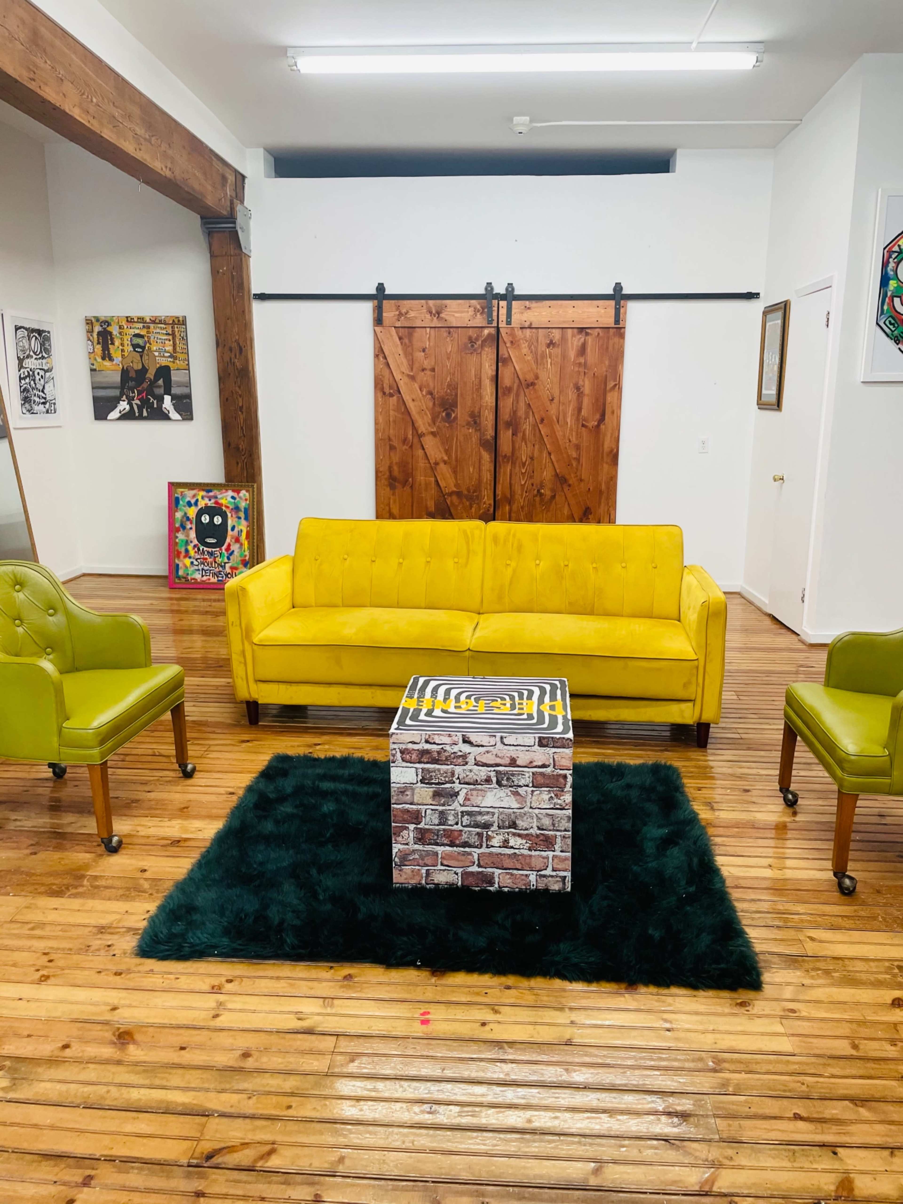 A bright yellow sofa surrounded by green chairs on a dark rug, with a brick-patterned coffee table in a spacious room featuring wooden floors and sliding wooden doors.