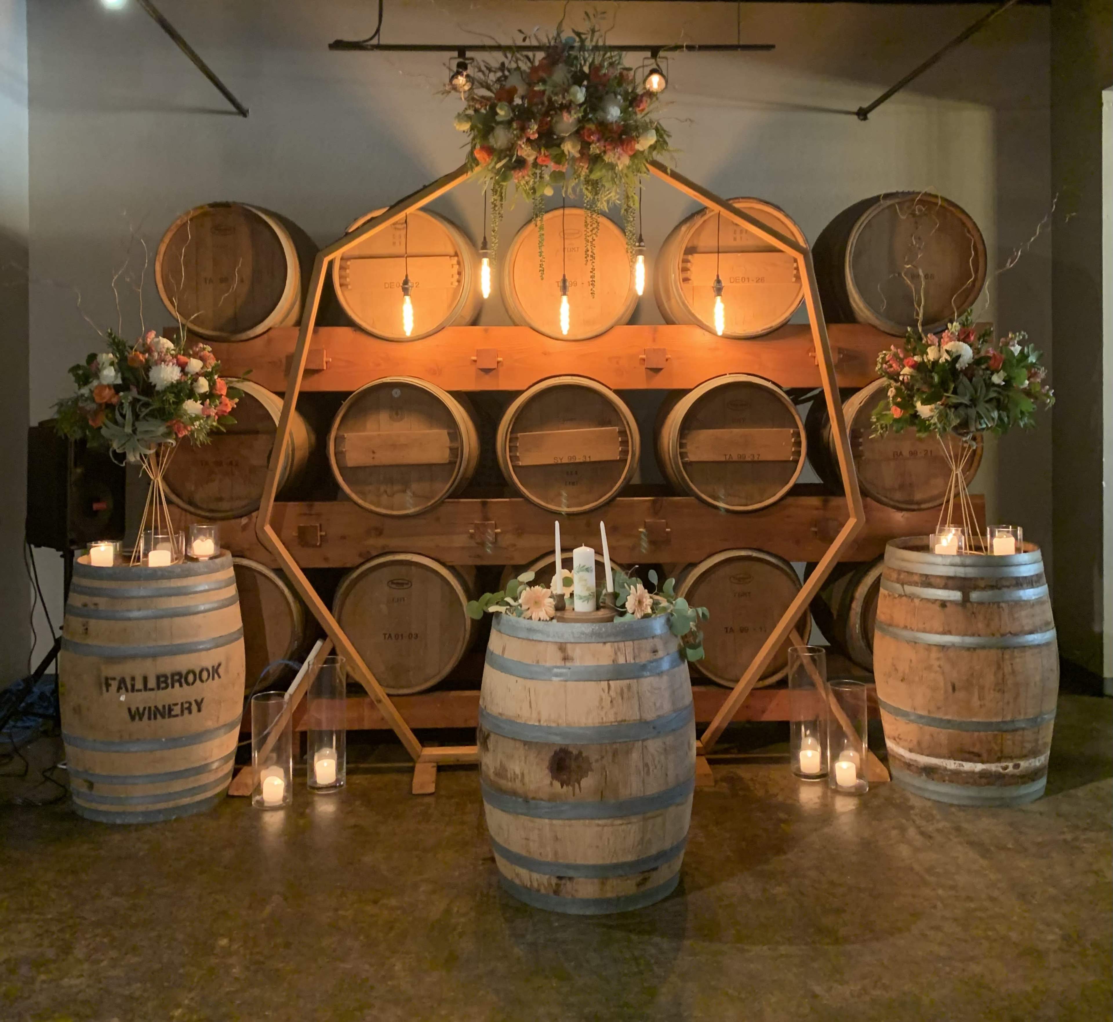 The scene features a wooden structure displaying wine barrels, adorned with floral arrangements, candles, and a centerpiece on a round barrel.