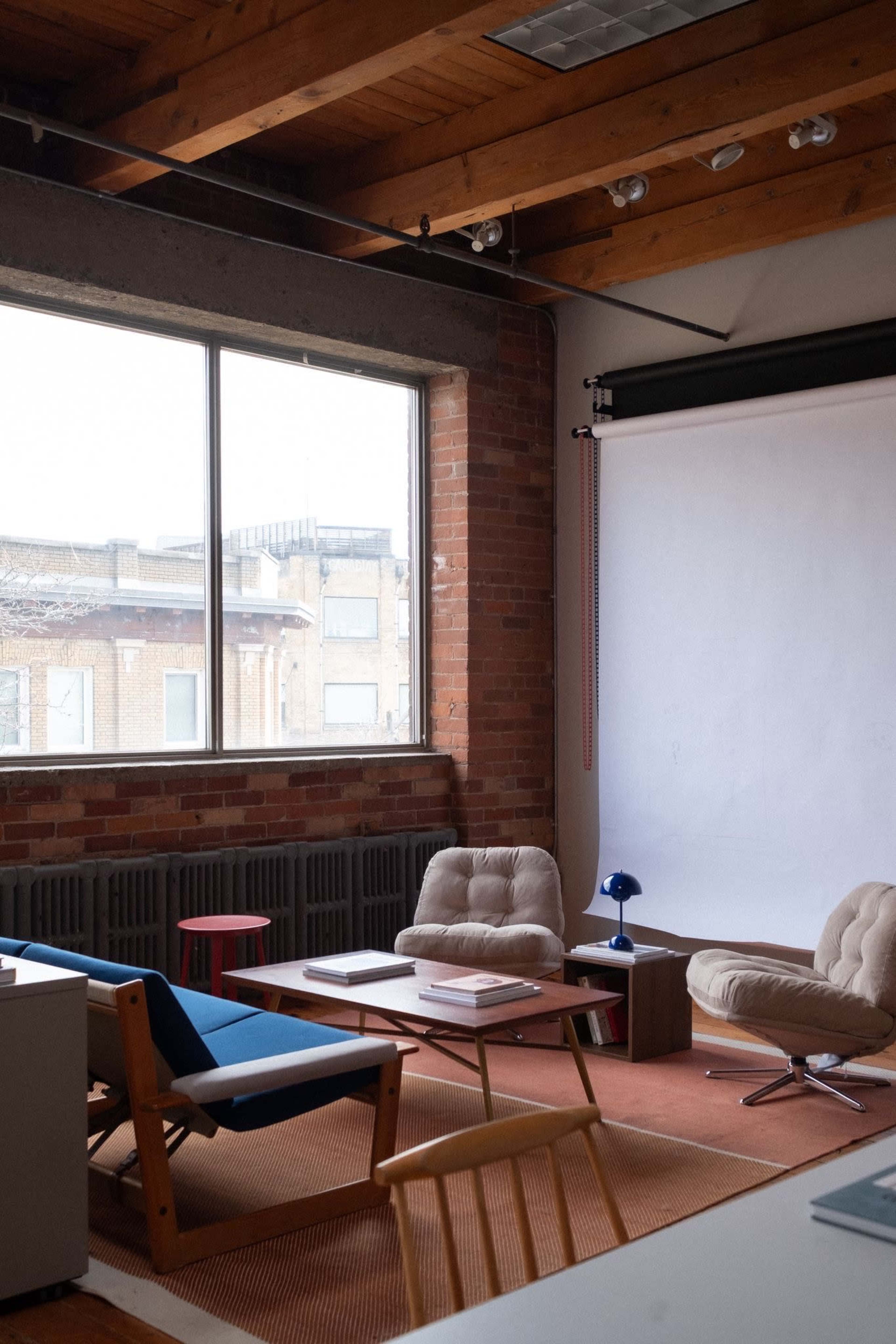 The image shows a modern room with brick walls, wooden beams, and minimalist furniture, including chairs and a table, beside a large window.