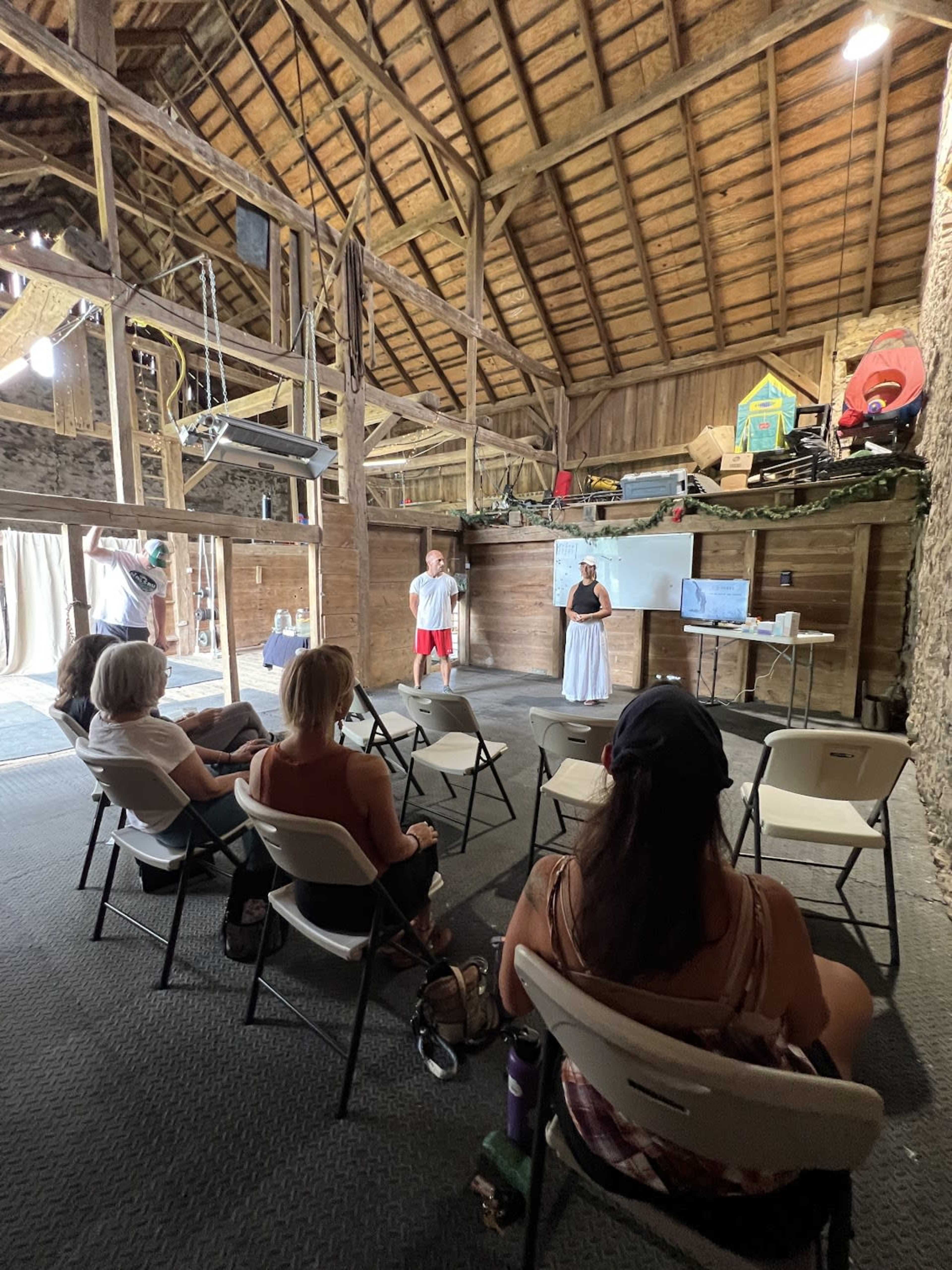 Two presenters stand in front of an audience seated in a barn, with a whiteboard and a projector displayed on a table.