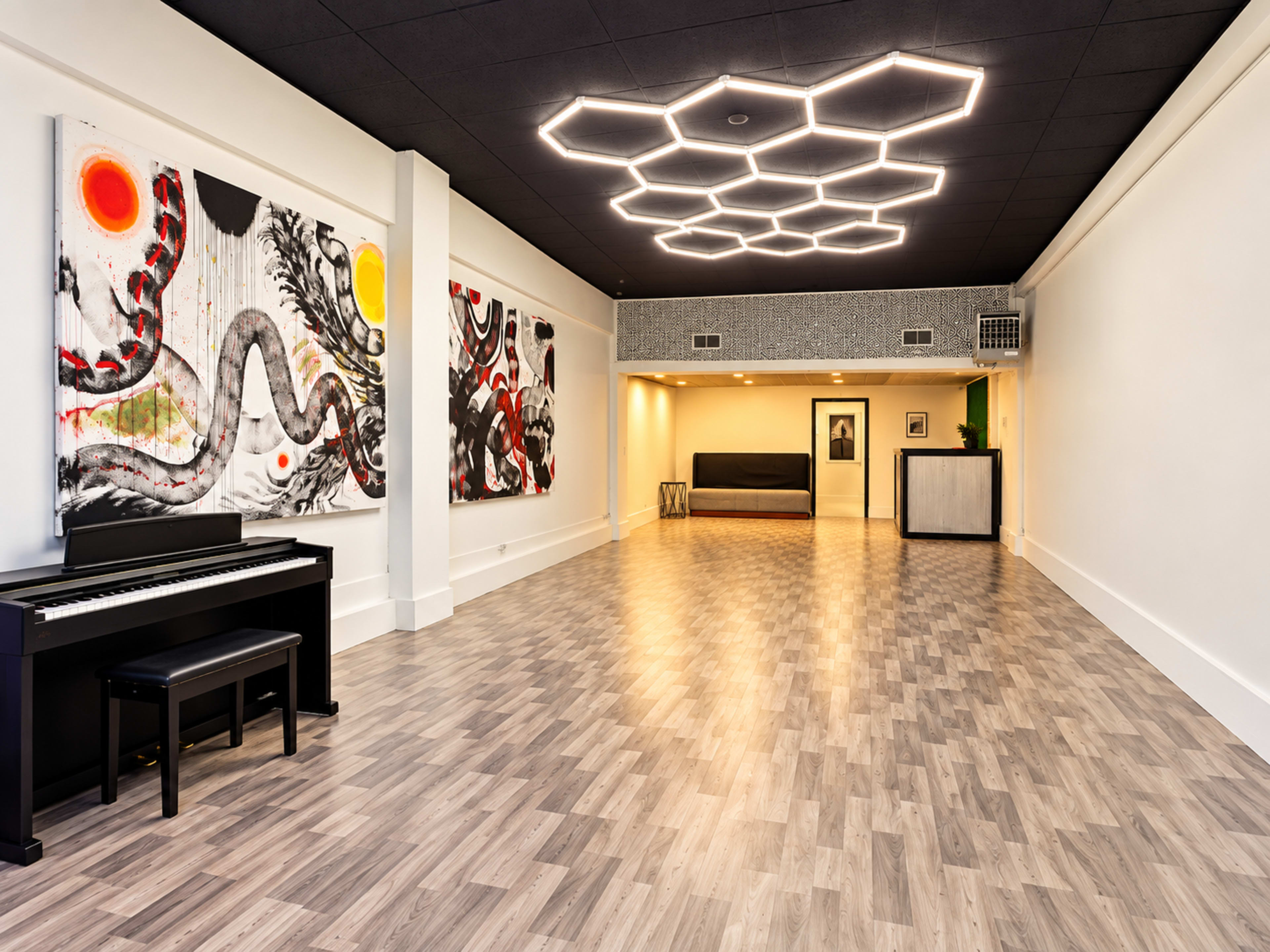 The image shows a modern, spacious interior with light-colored wooden flooring, abstract artwork on the walls, a black piano on the left, and a seating area in the background under a geometric light fixture.