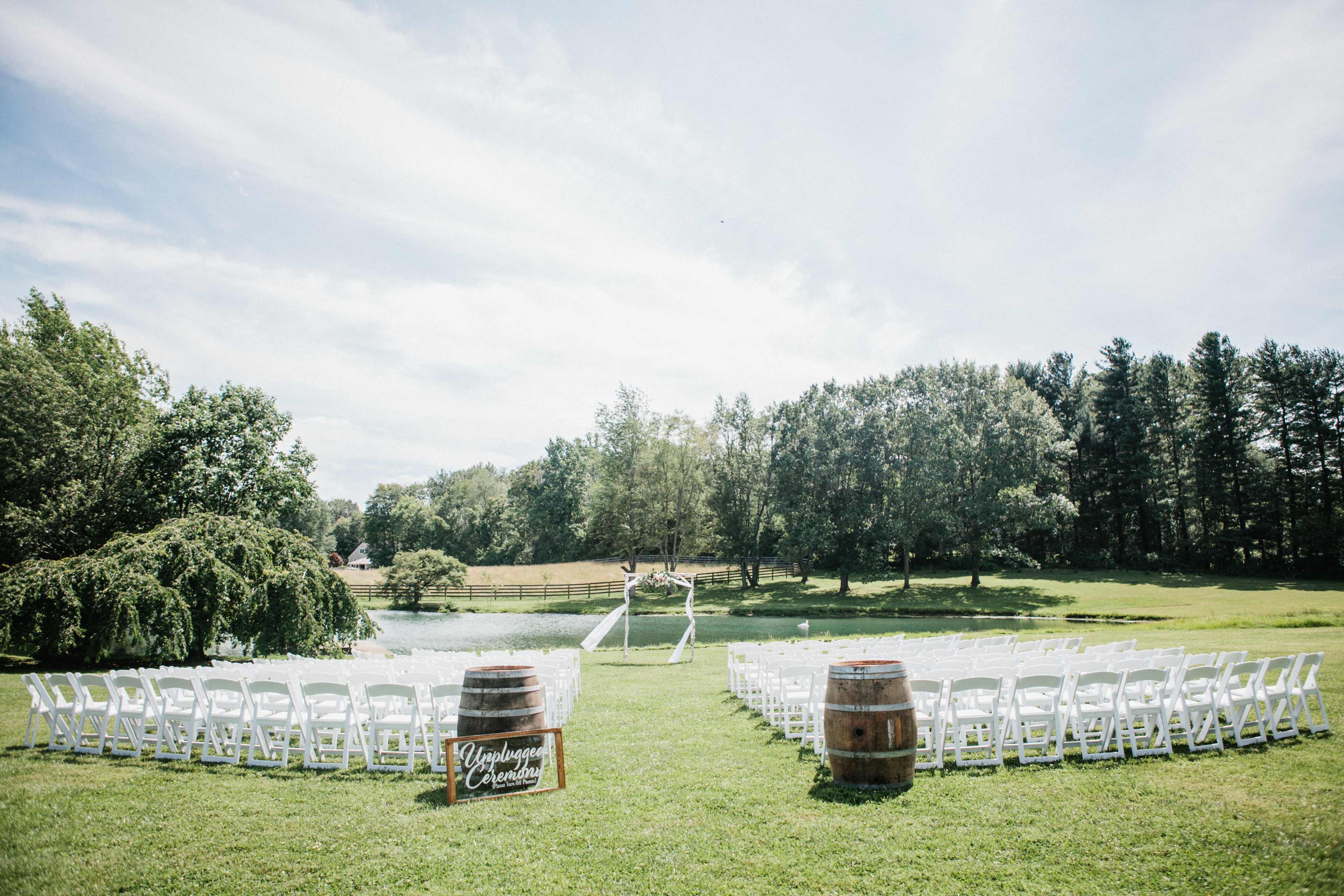 A wedding ceremony setup featuring rows of white chairs arranged on grass near a pond, with trees and a wooden arch in the background.