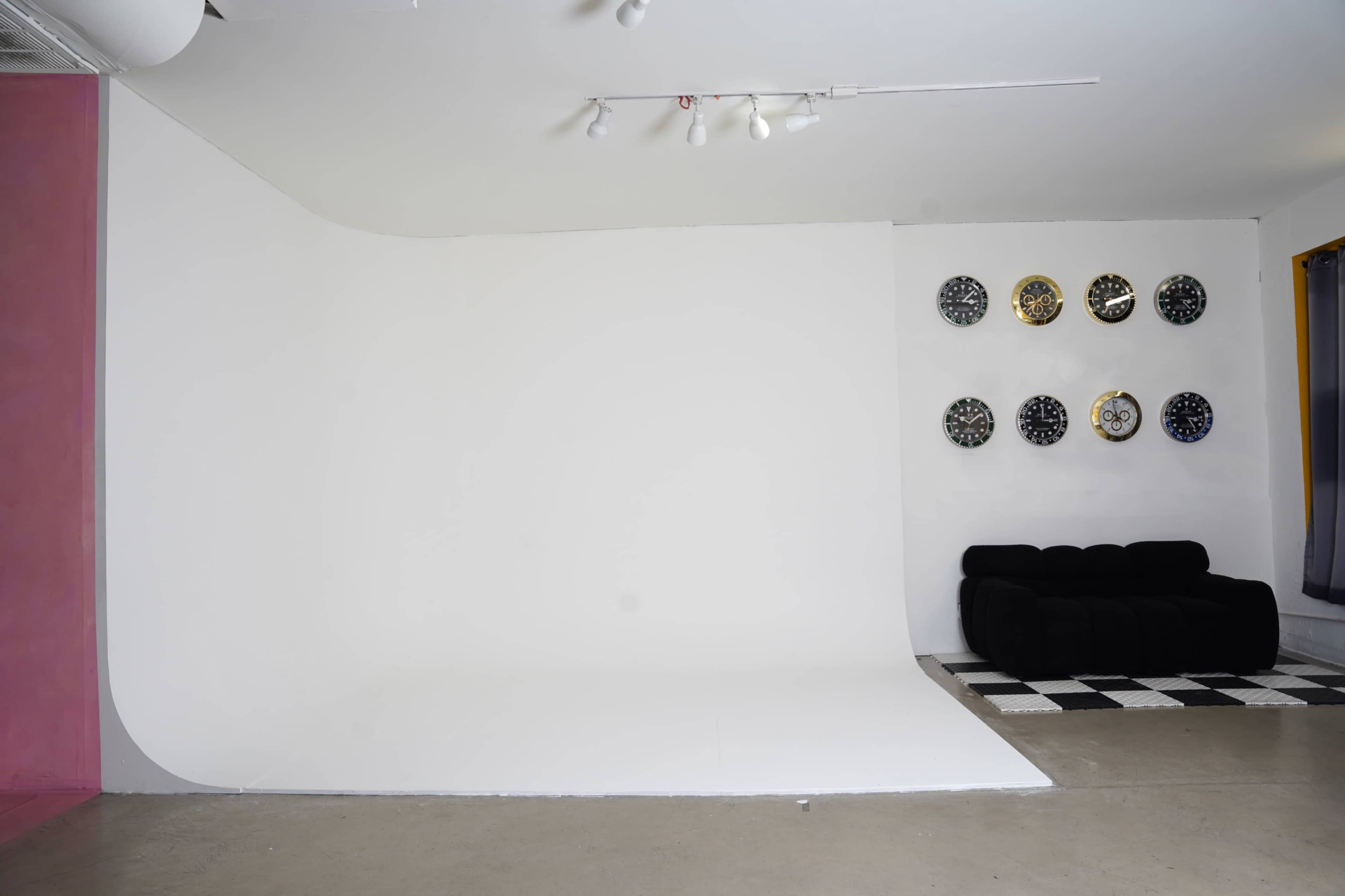 The image shows a minimalist studio space with a curved white backdrop, a black couch, and several decorative clocks mounted on the wall.