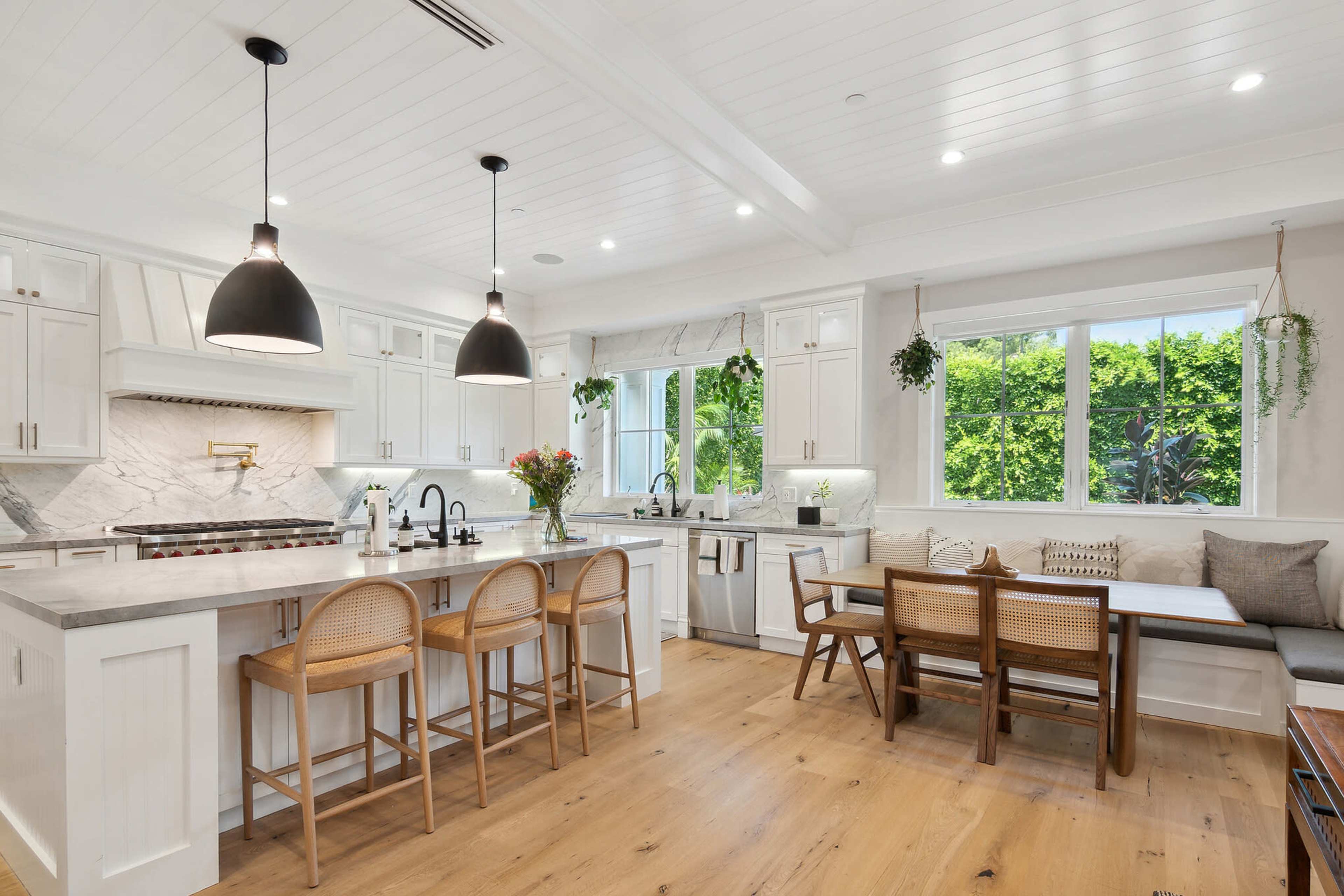A modern kitchen with white cabinetry, a marble backsplash, a large island with seating, and a dining area filled with natural light from a window.