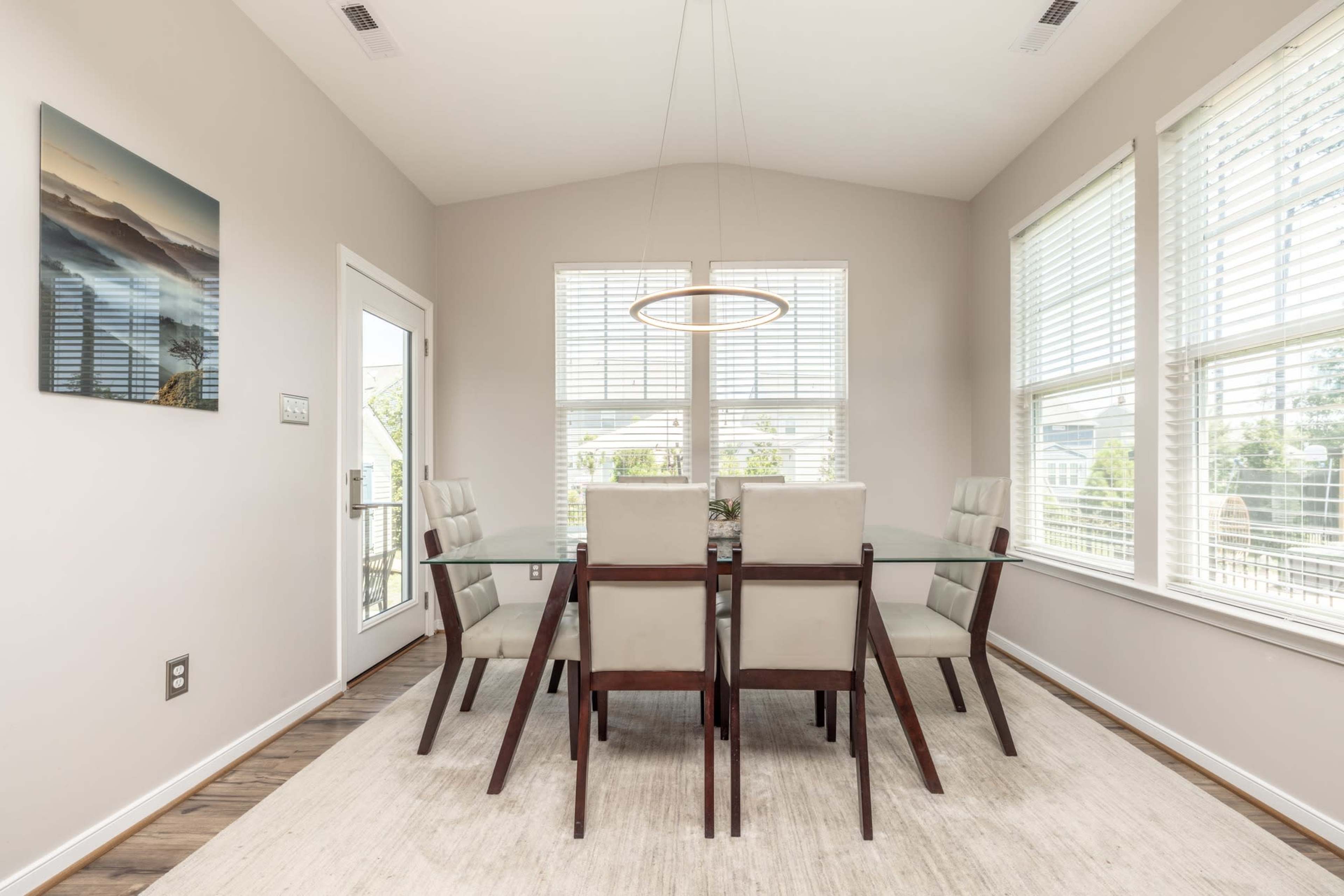 A modern dining room features a glass table surrounded by six upholstered chairs, with large windows allowing natural light to fill the space.