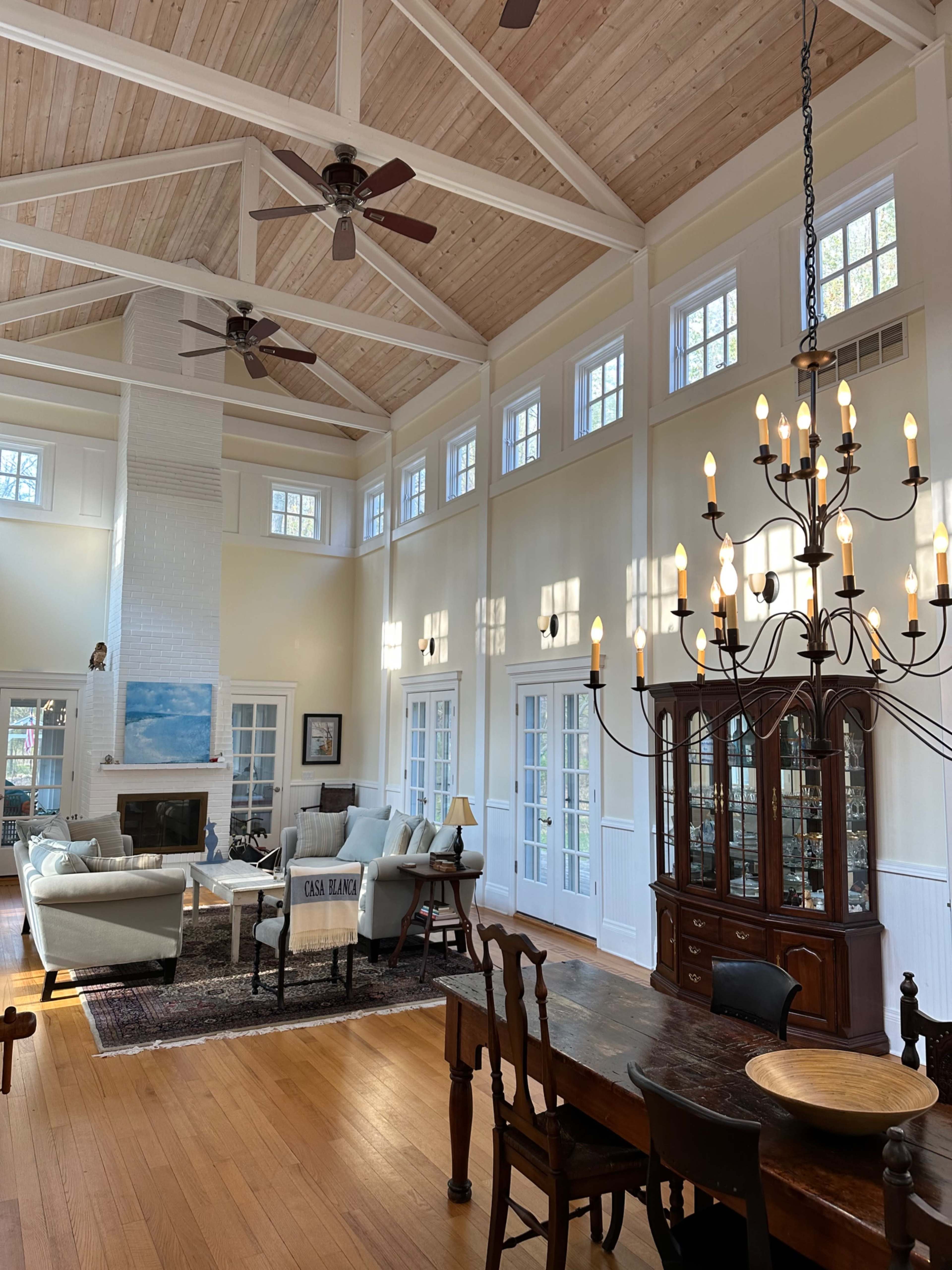 A spacious living area with vaulted ceilings, large windows, and a combination of a seating area and dining space, illuminated by a chandelier.