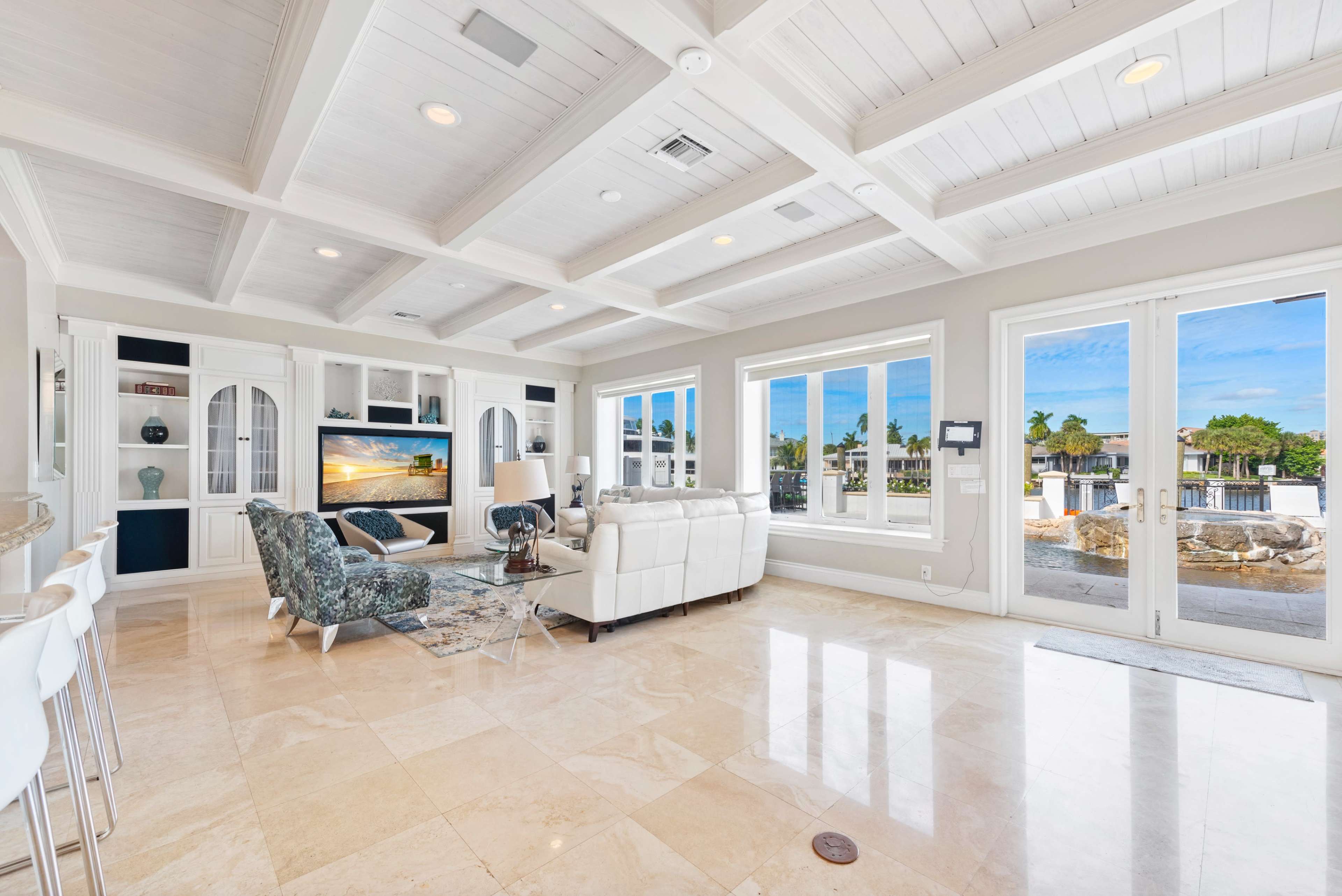 The image shows a modern living room with a white leather sofa, a patterned armchair, a coffee table, and large windows that offer a view of an outdoor area.