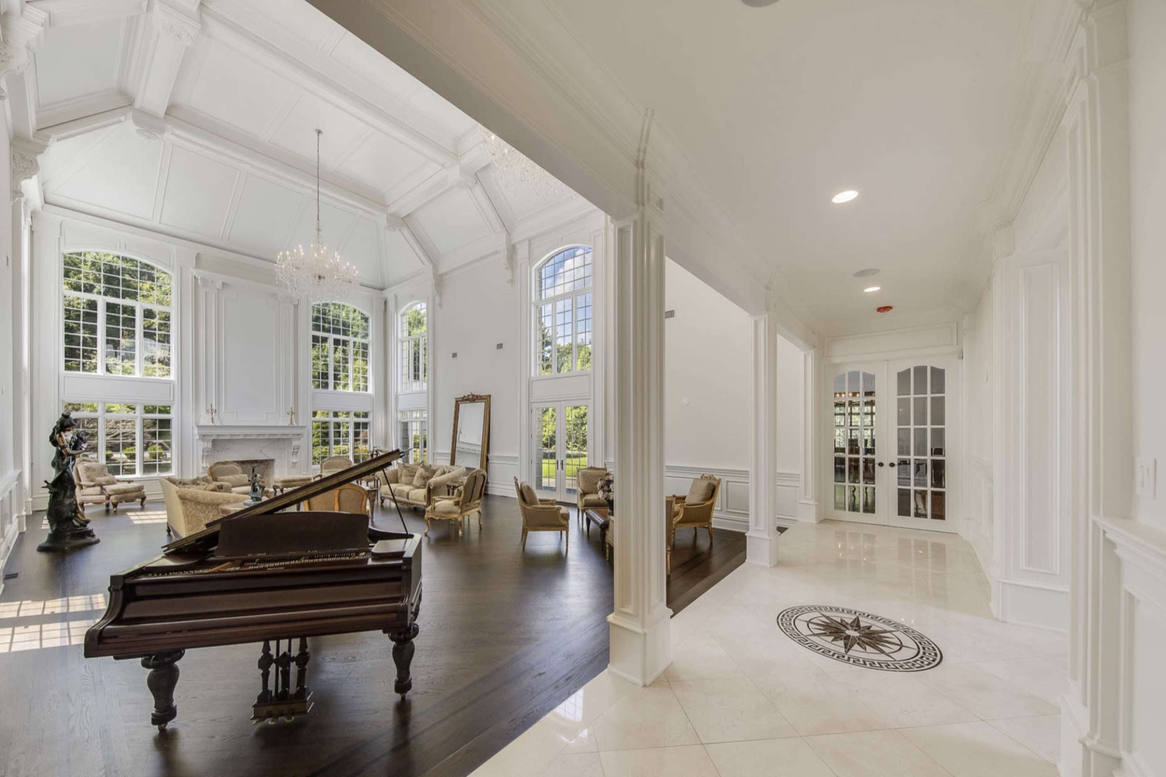 The image depicts a spacious, well-lit interior of a luxurious home featuring a grand piano, elegant seating areas, large windows, and intricate architectural details.