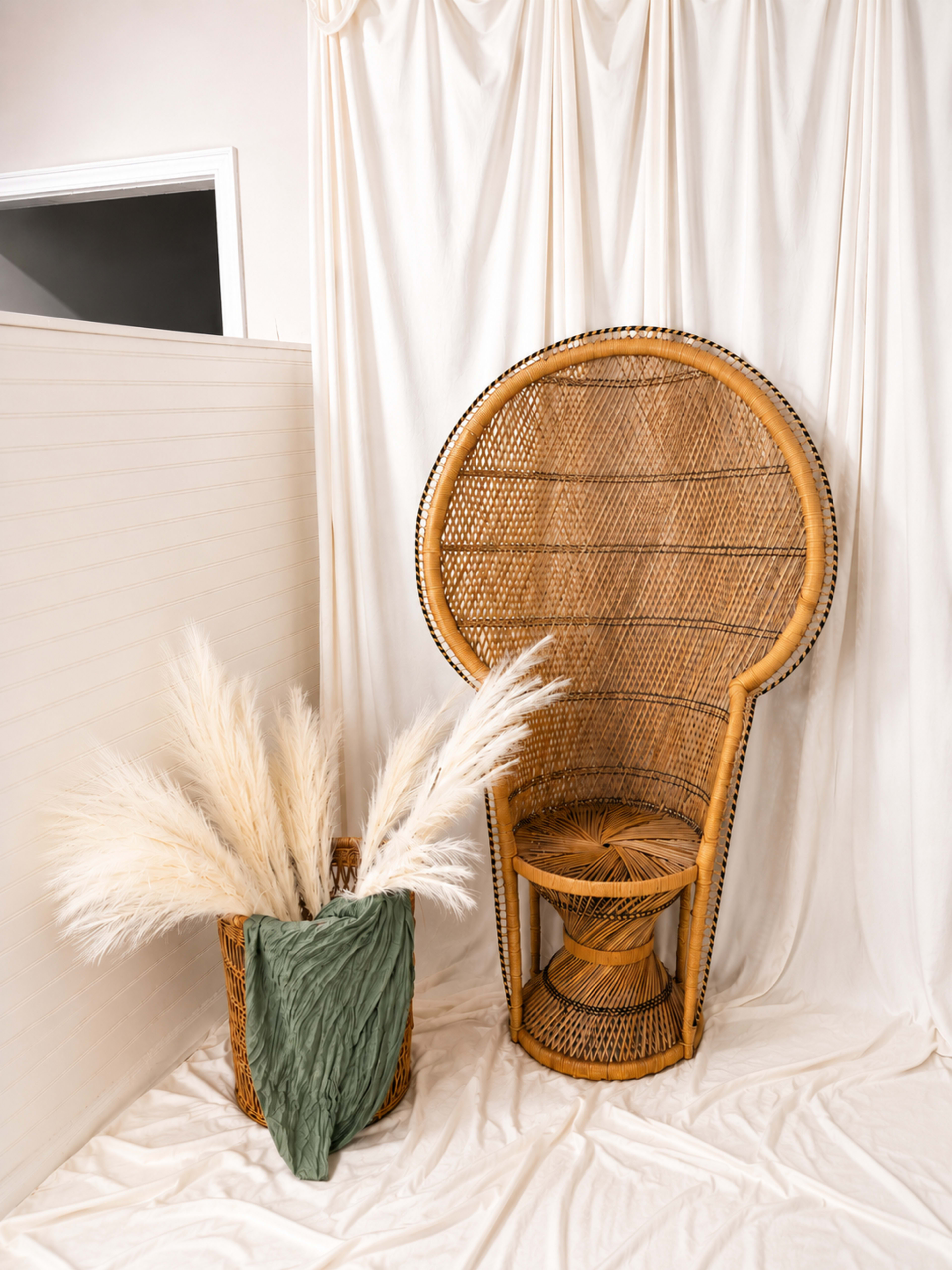 A large, woven rattan chair is positioned beside a basket filled with pampas grass and a green fabric draped over the side, set against a backdrop of white drapery.