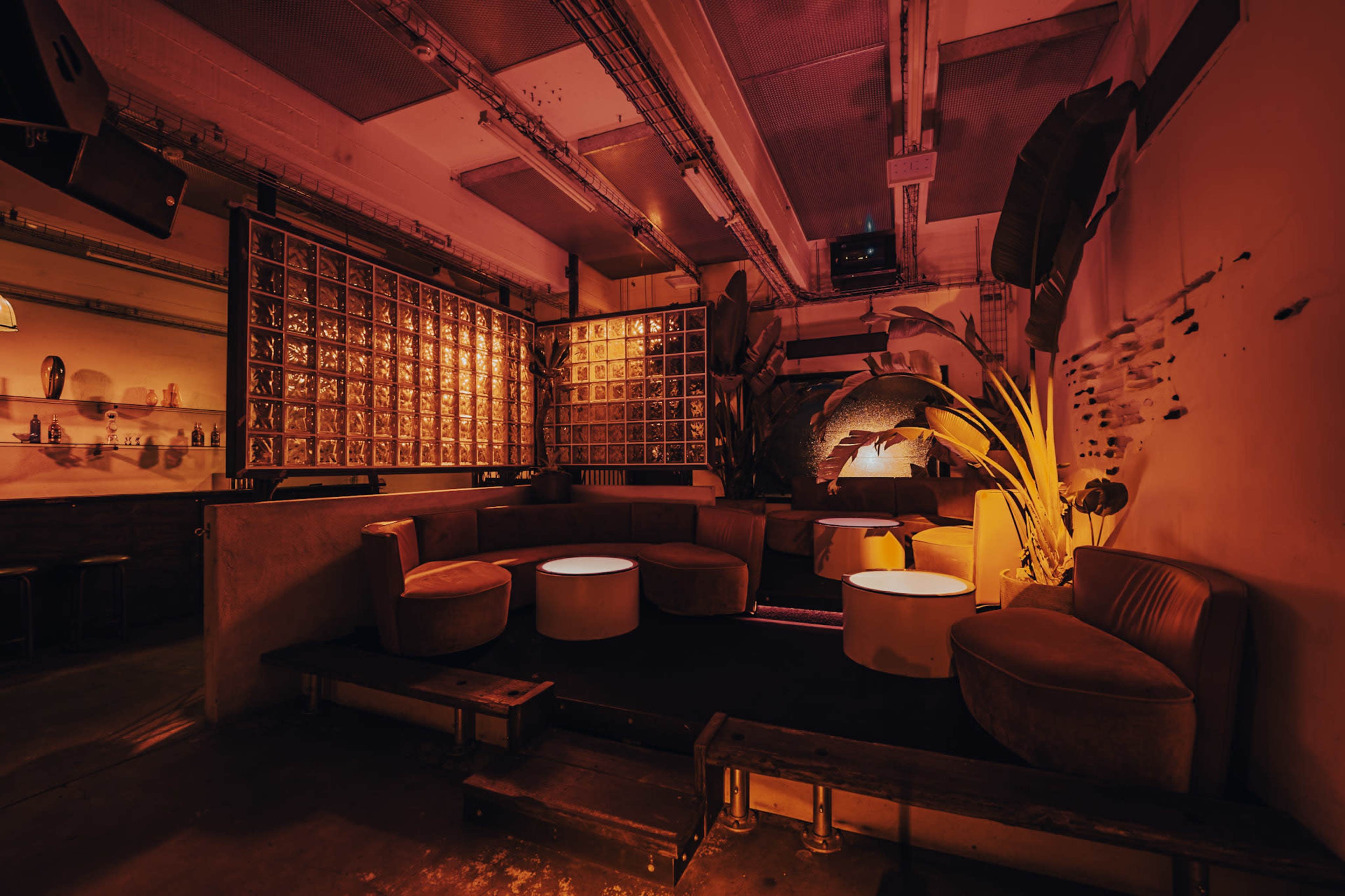 The image shows a dimly lit lounge area featuring circular seating, low tables, and a backdrop of decorative panels, all illuminated by warm orange light.