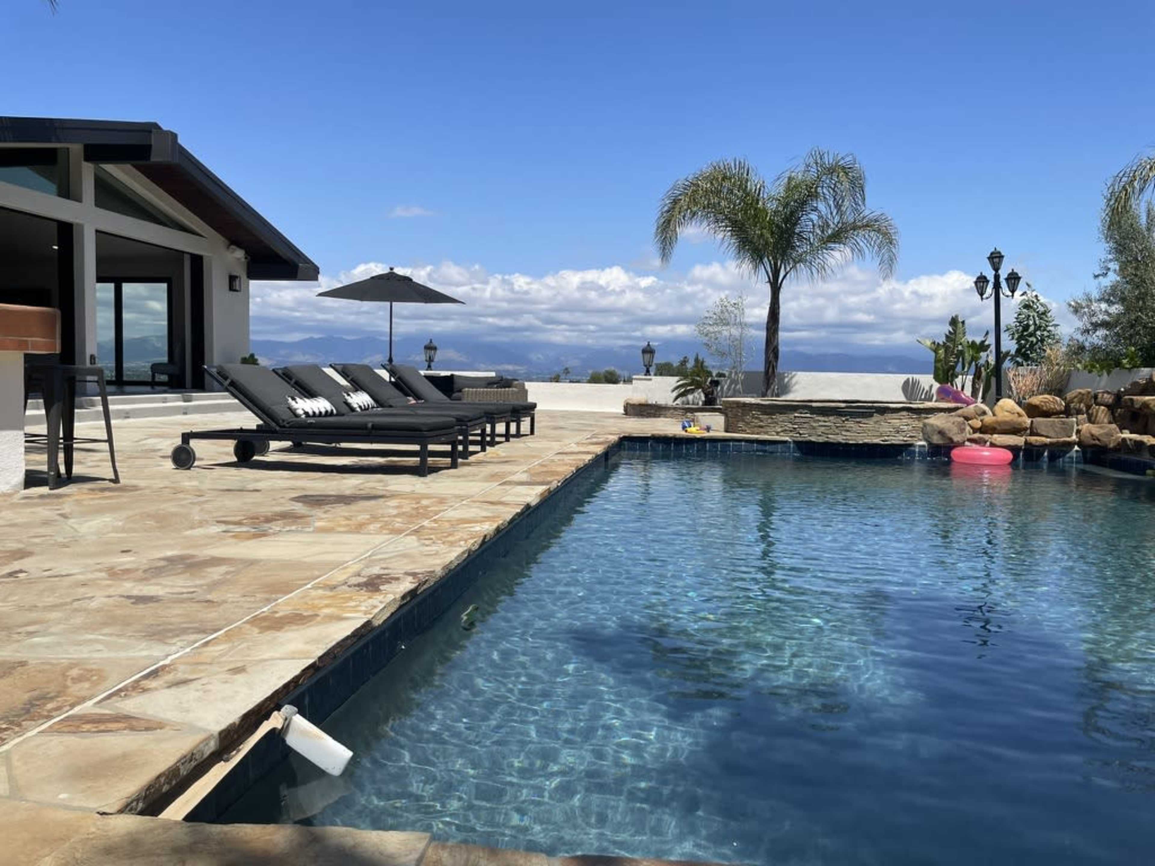 A swimming pool with lounge chairs is set on a stone patio, overlooking a landscape with palm trees and distant mountains under a blue sky.