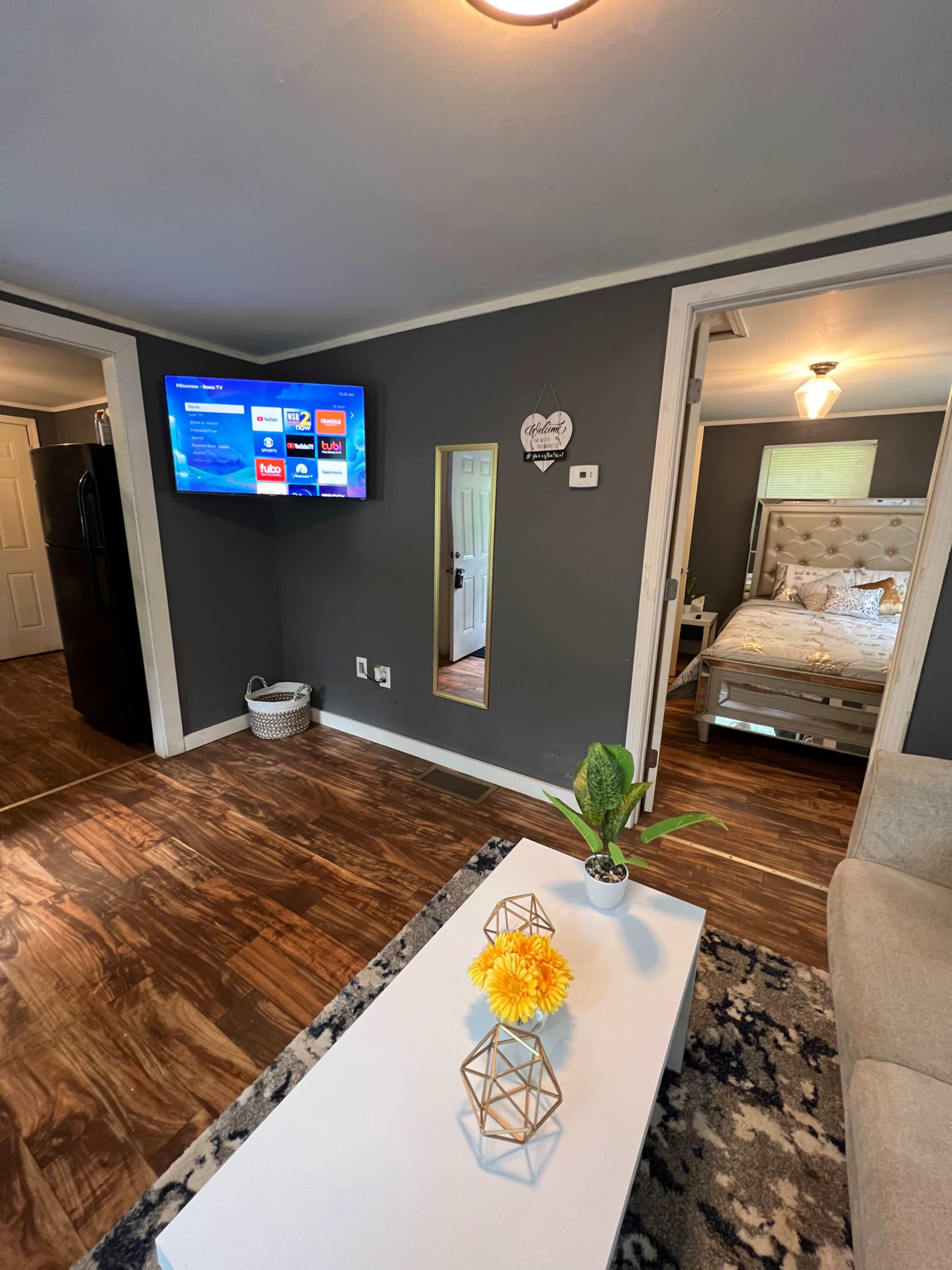 A living room with a television mounted on the wall, a coffee table with flowers, and a mirror reflecting a bedroom doorway.