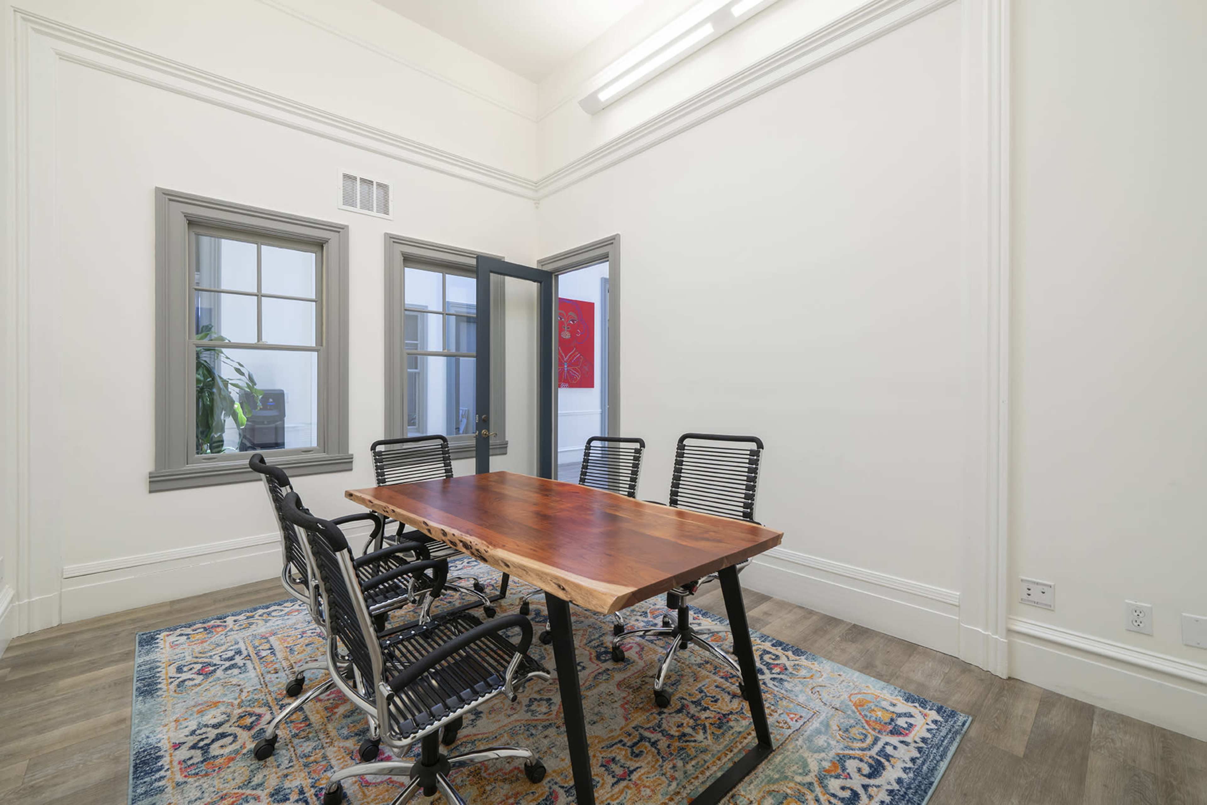 A conference room features a wooden table surrounded by five black office chairs, with large windows and a patterned rug on the floor.