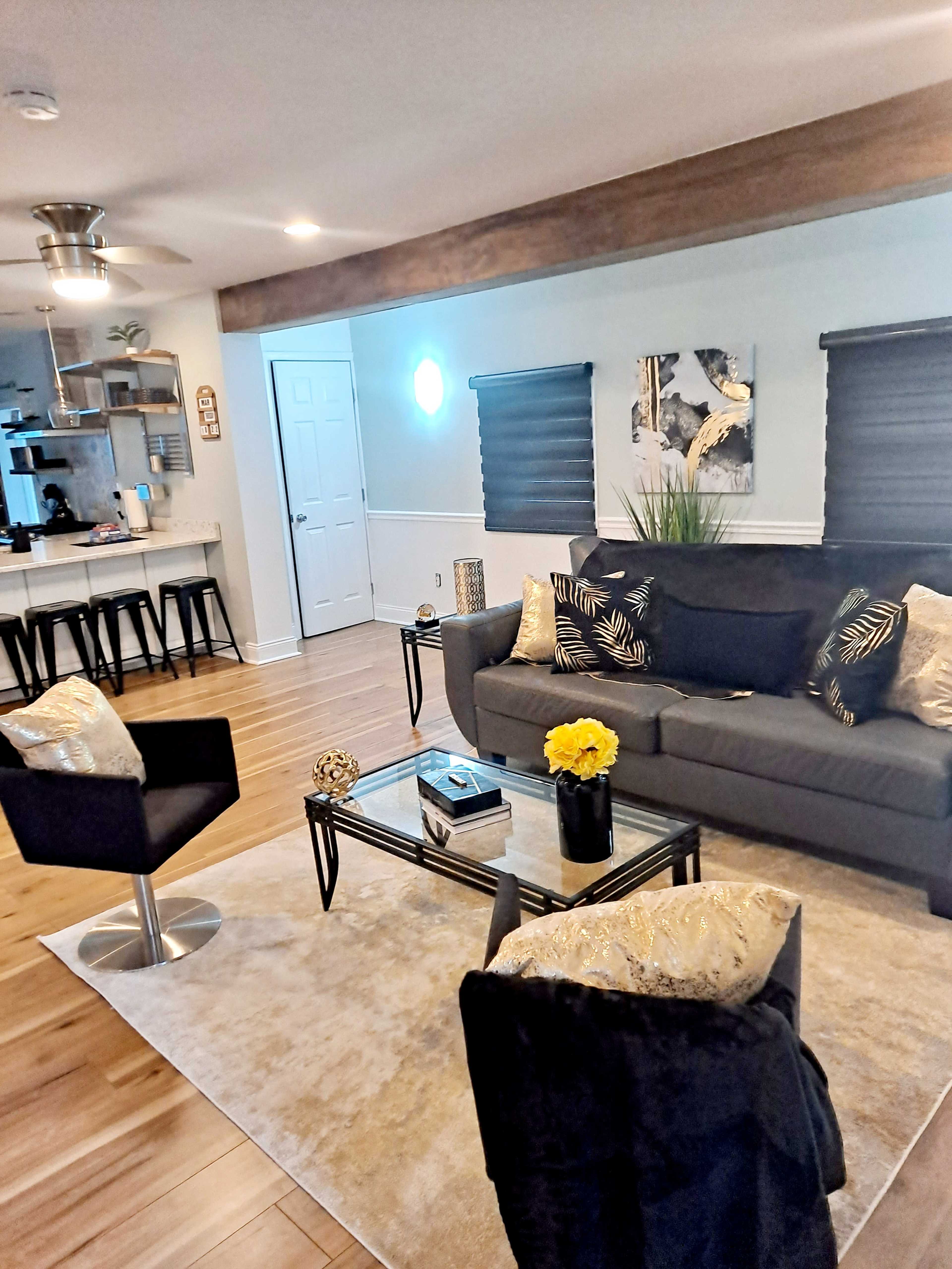 A modern living room features a gray sofa, black chairs, a glass coffee table, and a decorative rug, with open sightlines to a kitchen area.