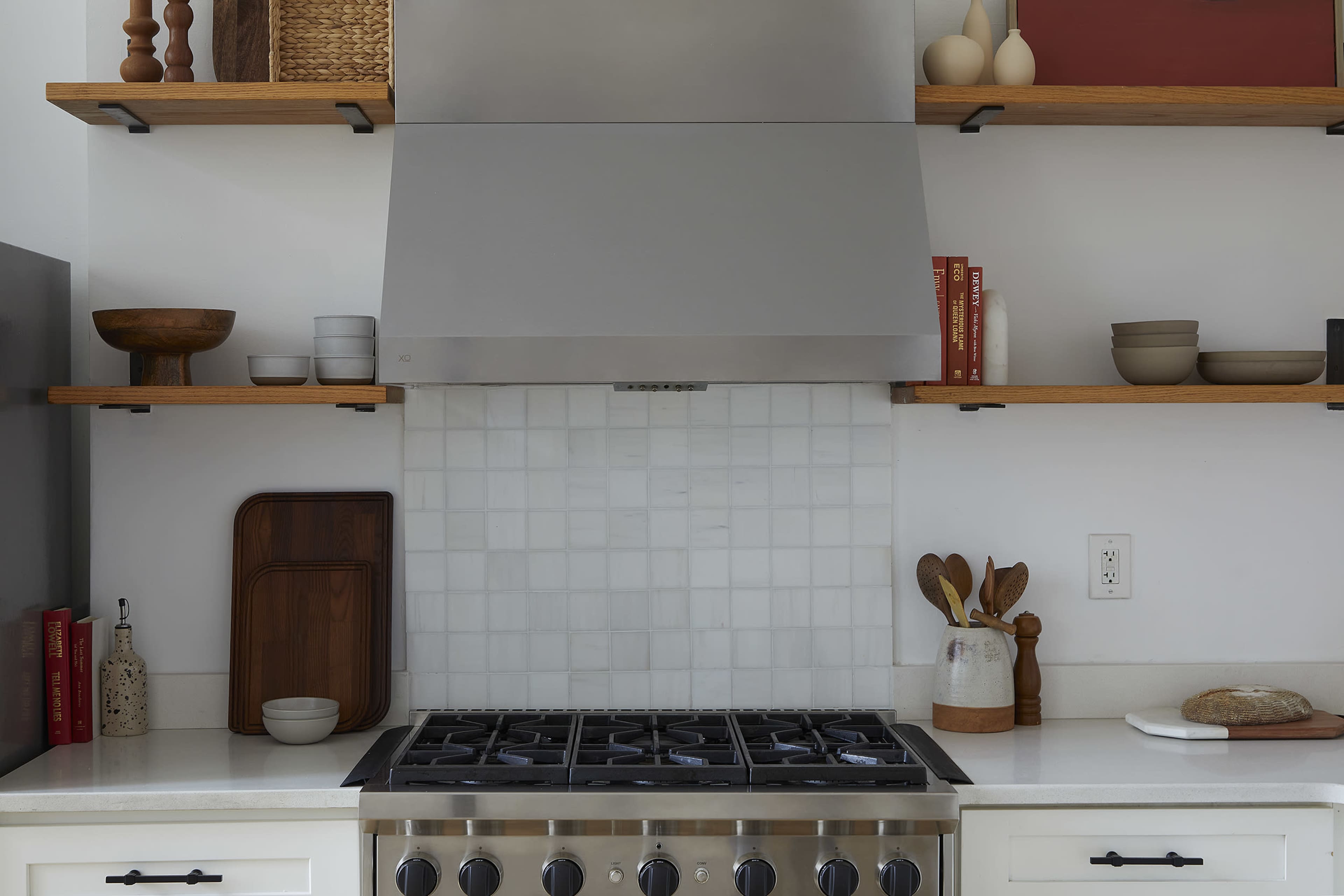 A modern kitchen features a stainless steel range hood above a gas stove, with open wooden shelves displaying various kitchenware and decor.