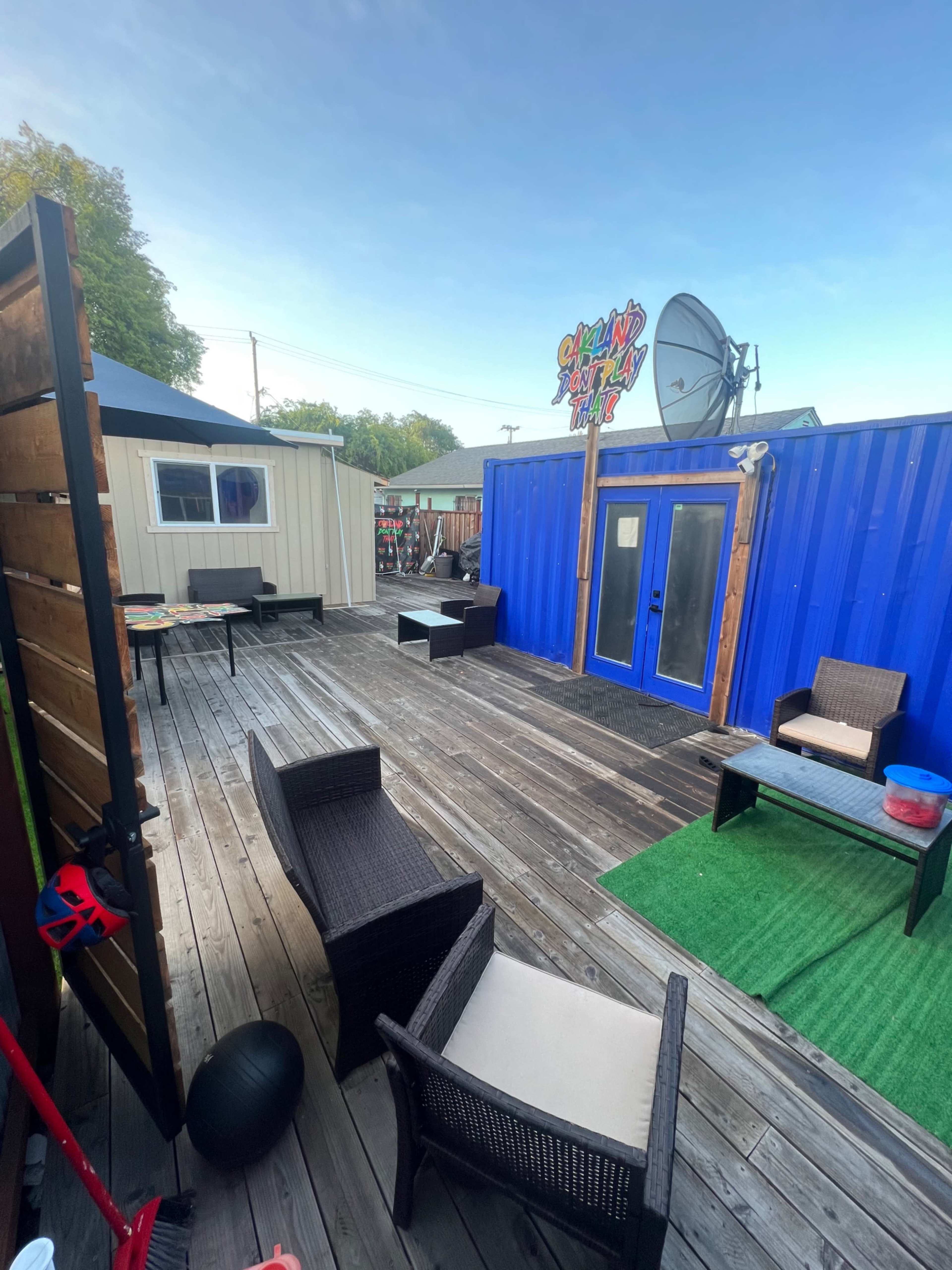 The image shows a wooden deck with several outdoor seating arrangements, including a table and chairs, surrounded by colorful buildings and a large satellite dish.