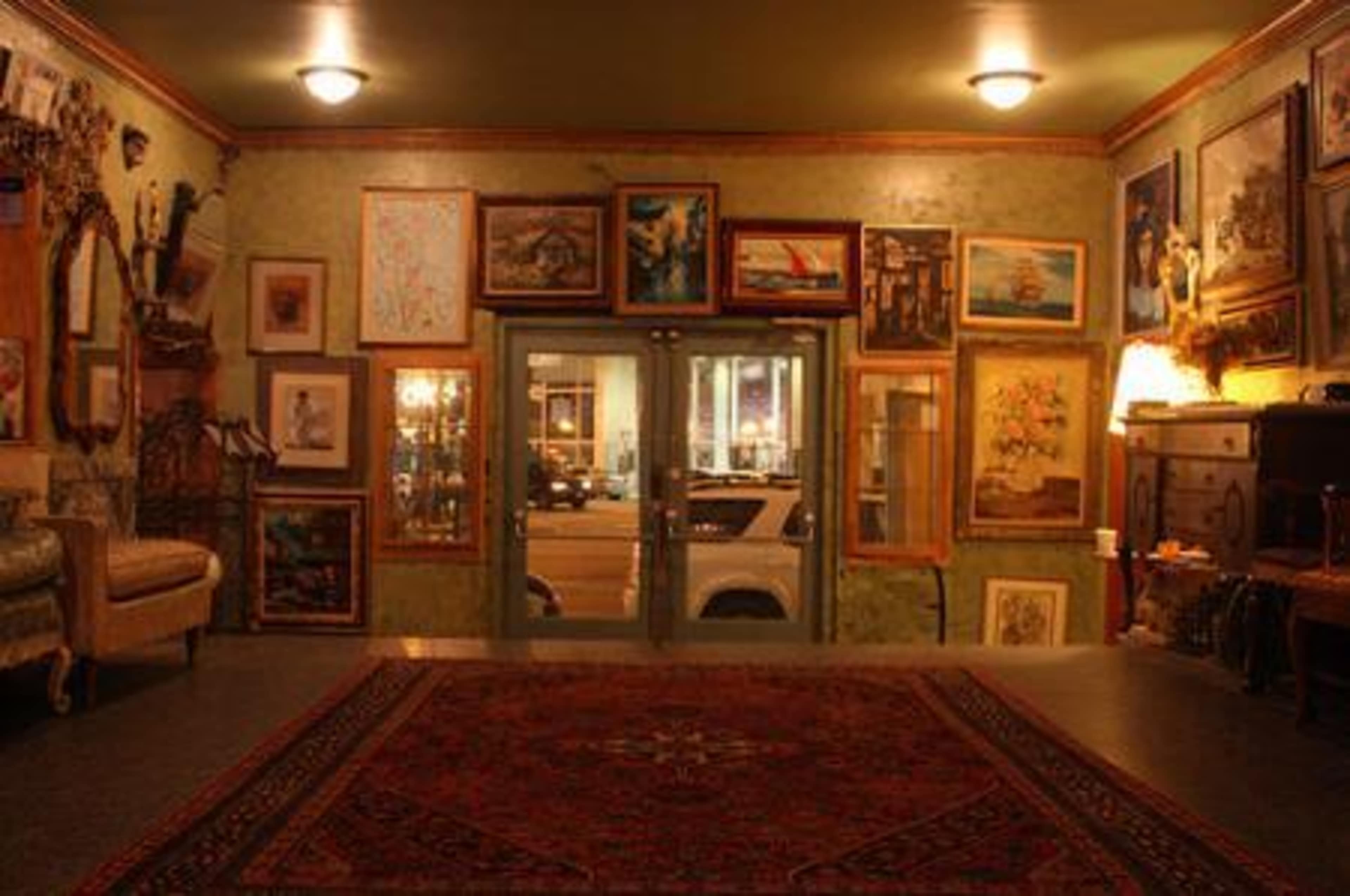 The image shows an art gallery interior with various framed paintings on the walls and a patterned rug on the floor.