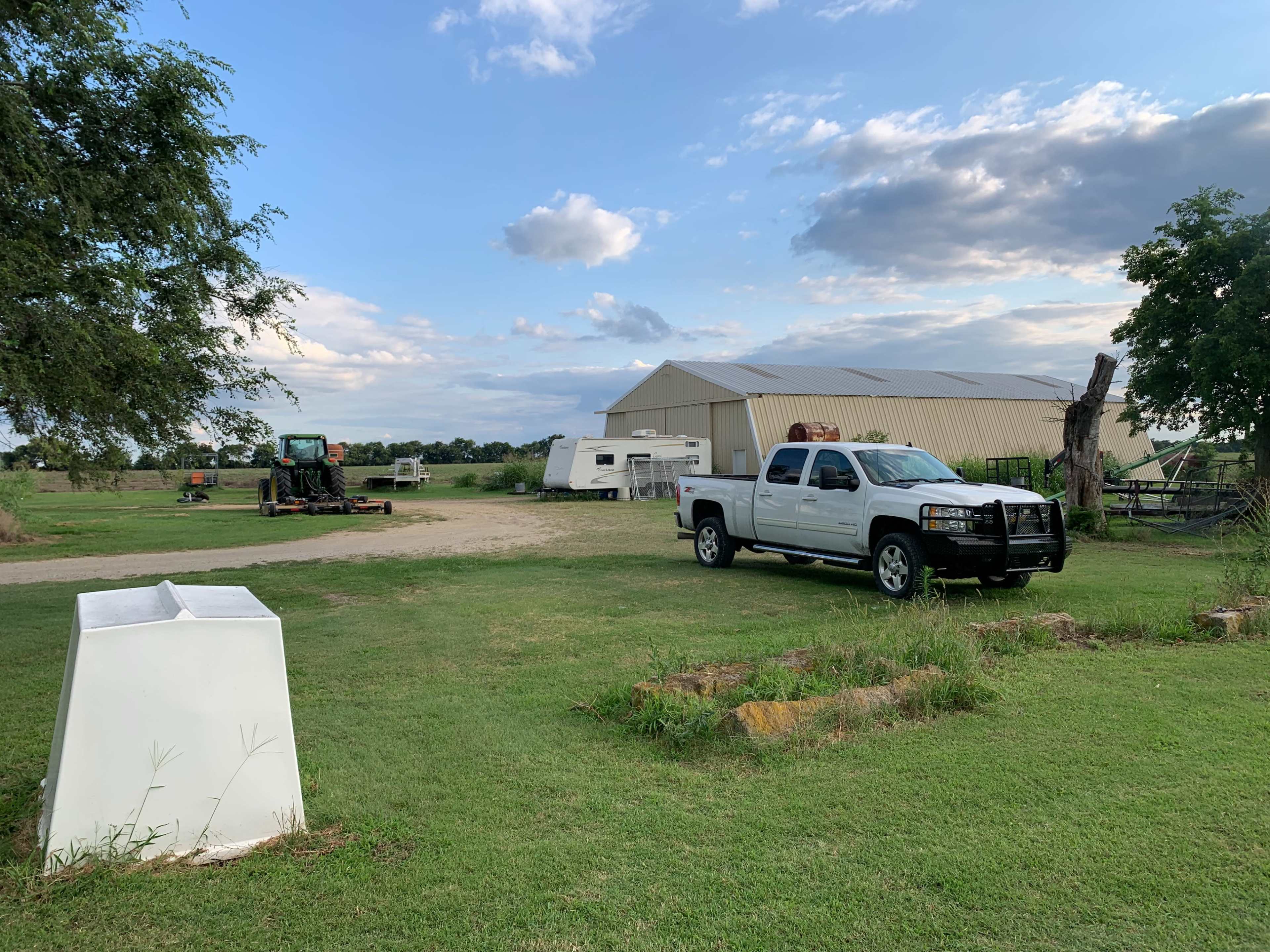 A white pickup truck is parked on a grassy area near a barn and parked trailers under a partly cloudy sky.
