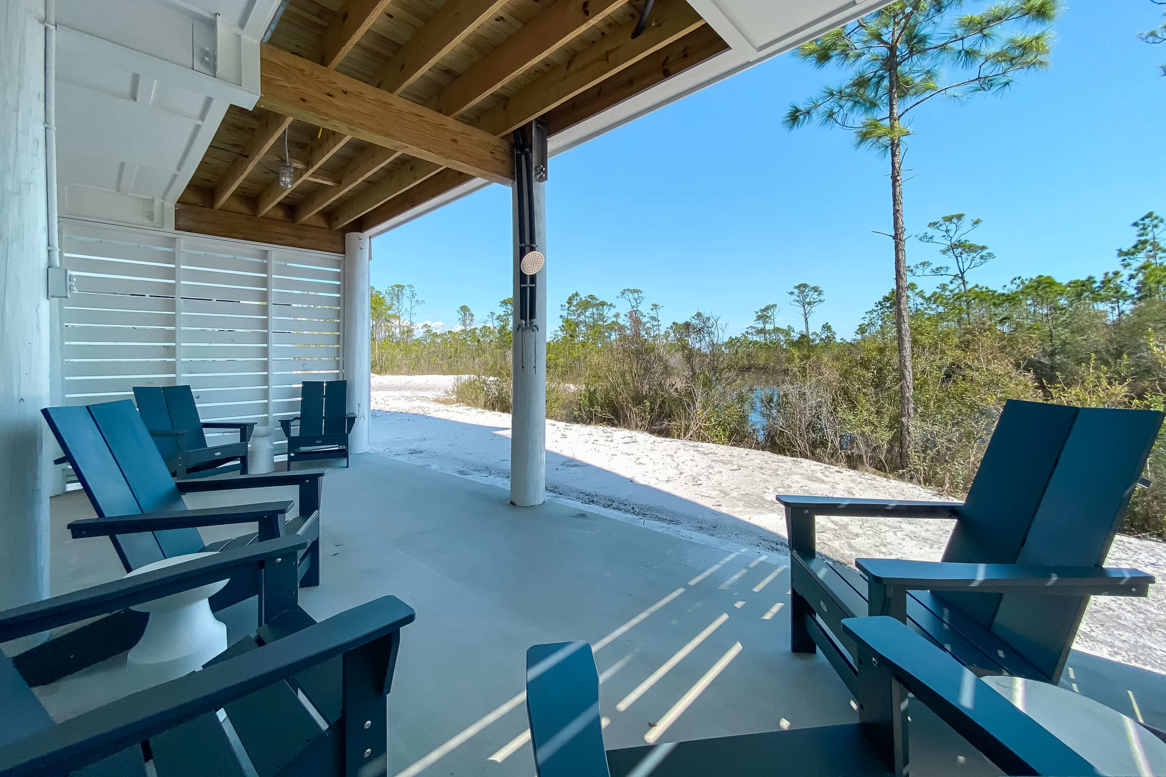 A covered patio with blue Adirondack chairs overlooks a sandy area and a body of water surrounded by vegetation.
