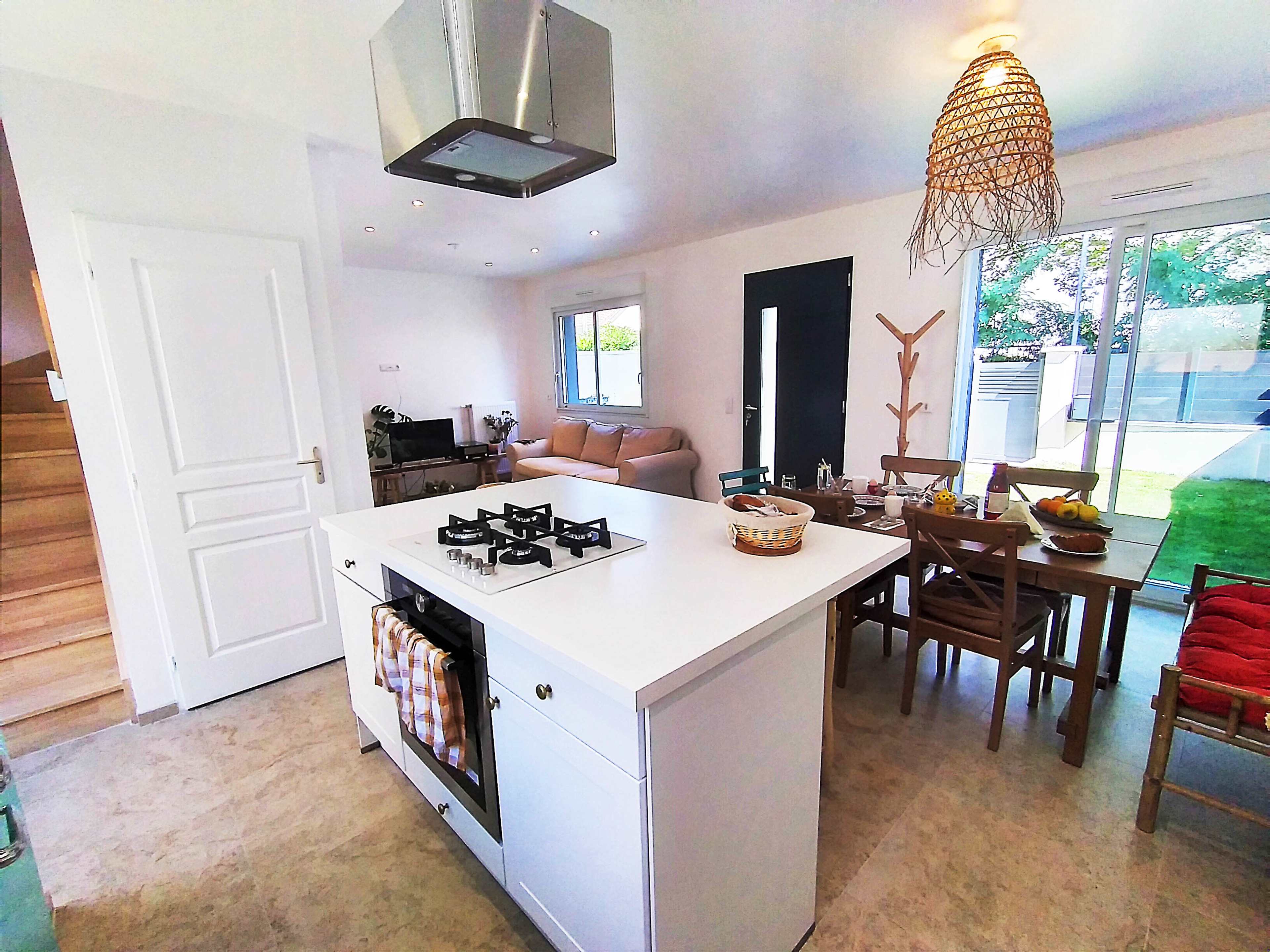 A modern kitchen with a central island, a gas stove, a dining area with wooden chairs, and large windows overlooking a garden.