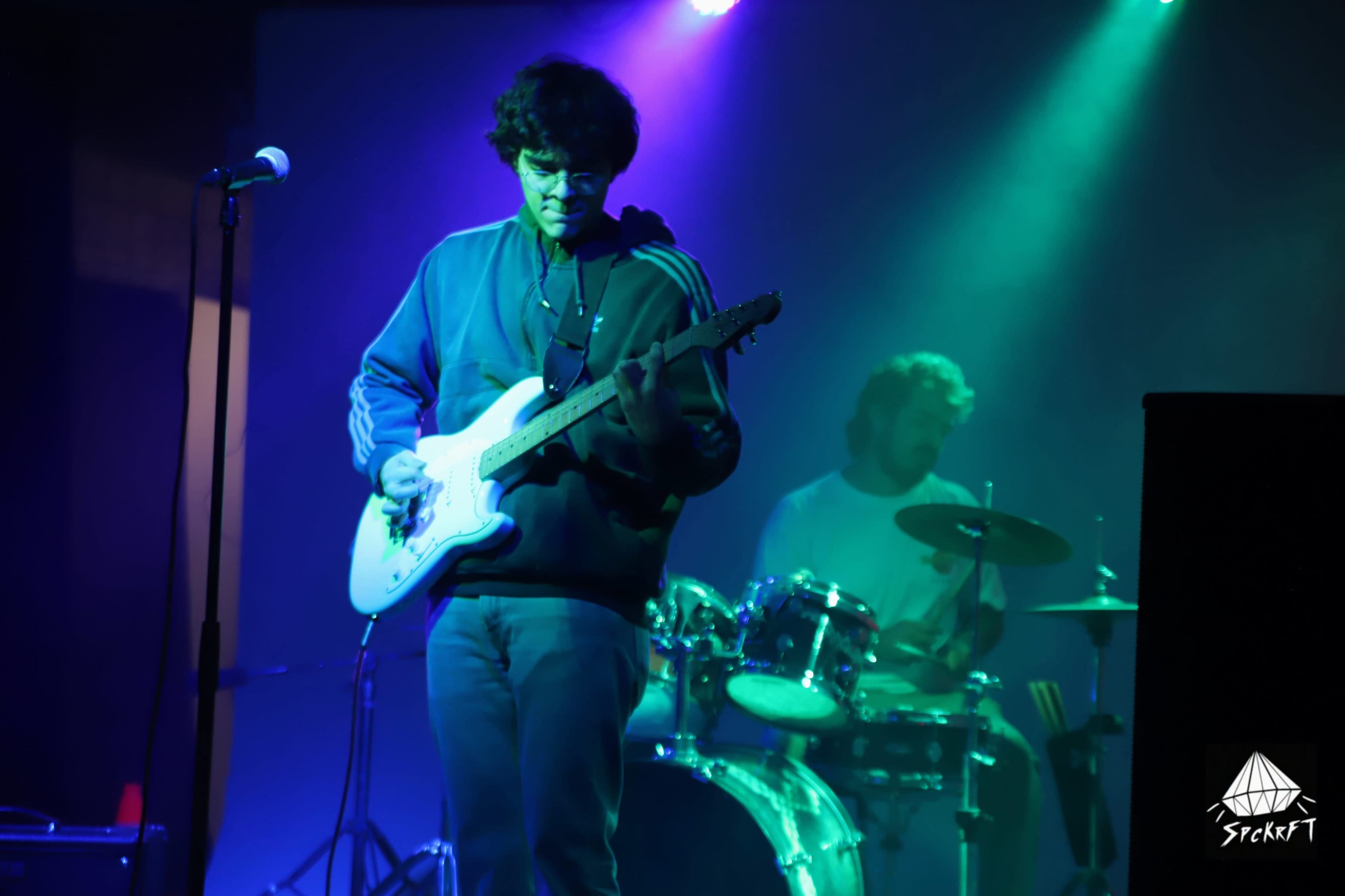 A musician plays electric guitar on stage while a drummer performs in the background, illuminated by colorful stage lights.