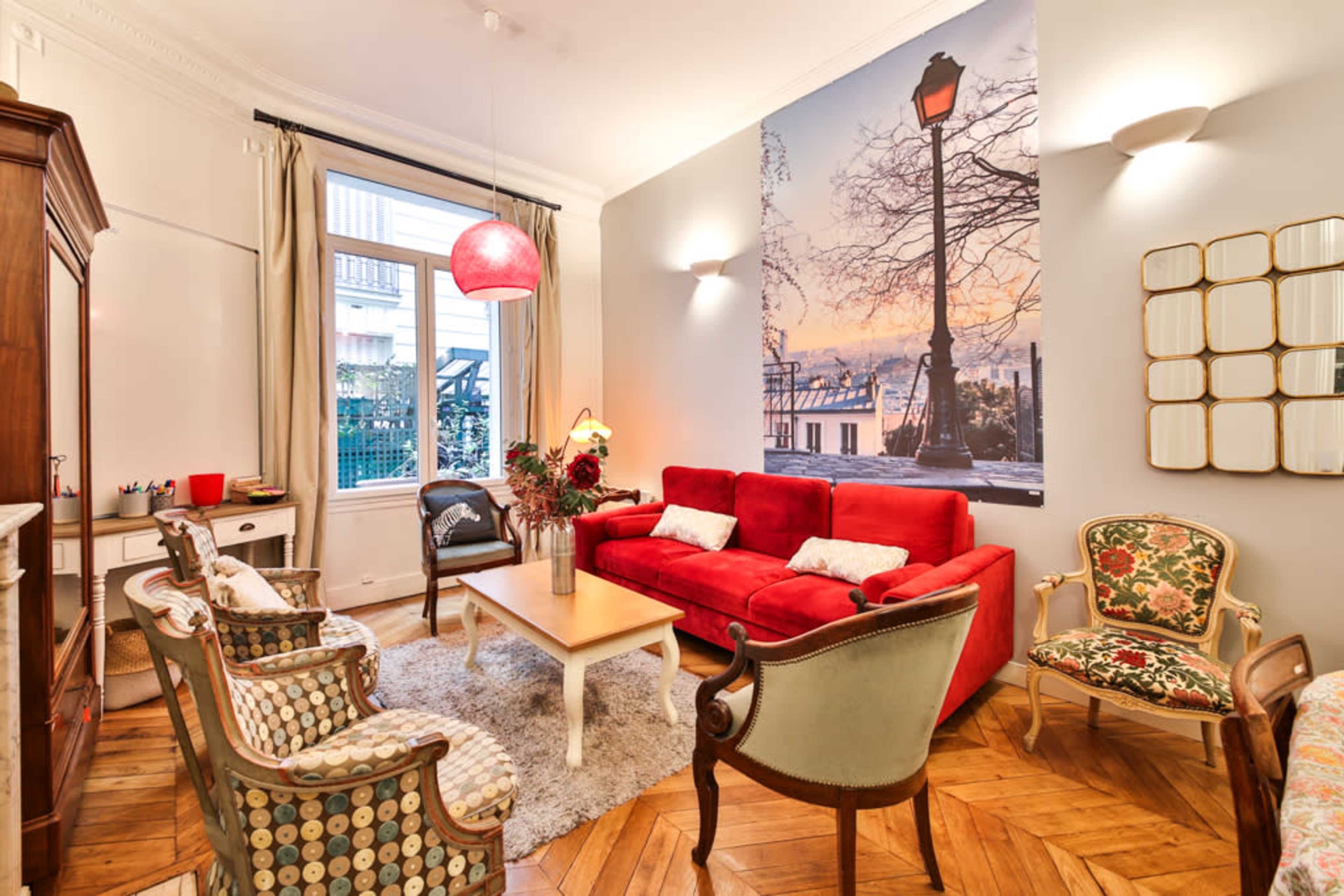 The image shows a cozy living room with a red couch, patterned chairs, a wooden cabinet, and a large wall mural of a sunset beside a lamppost.