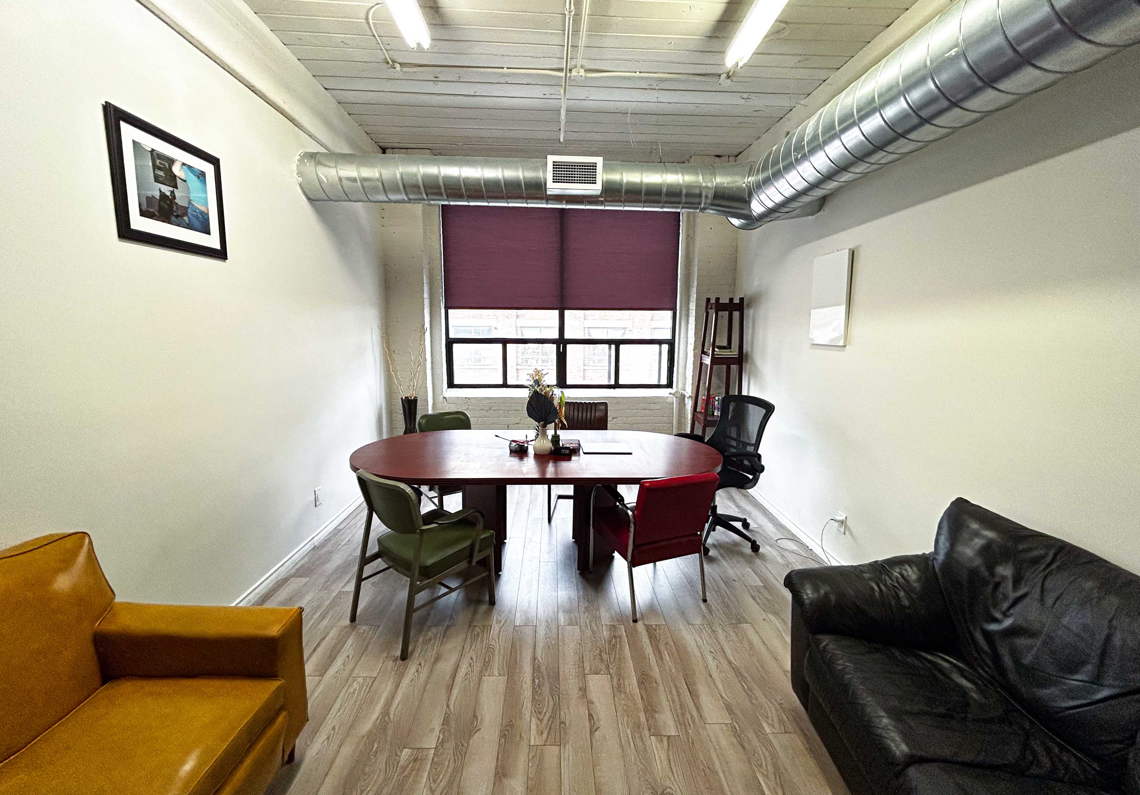 The image shows a simple office space featuring a round wooden table surrounded by chairs, a modern chair in the corner, and an exposed duct running along the ceiling.