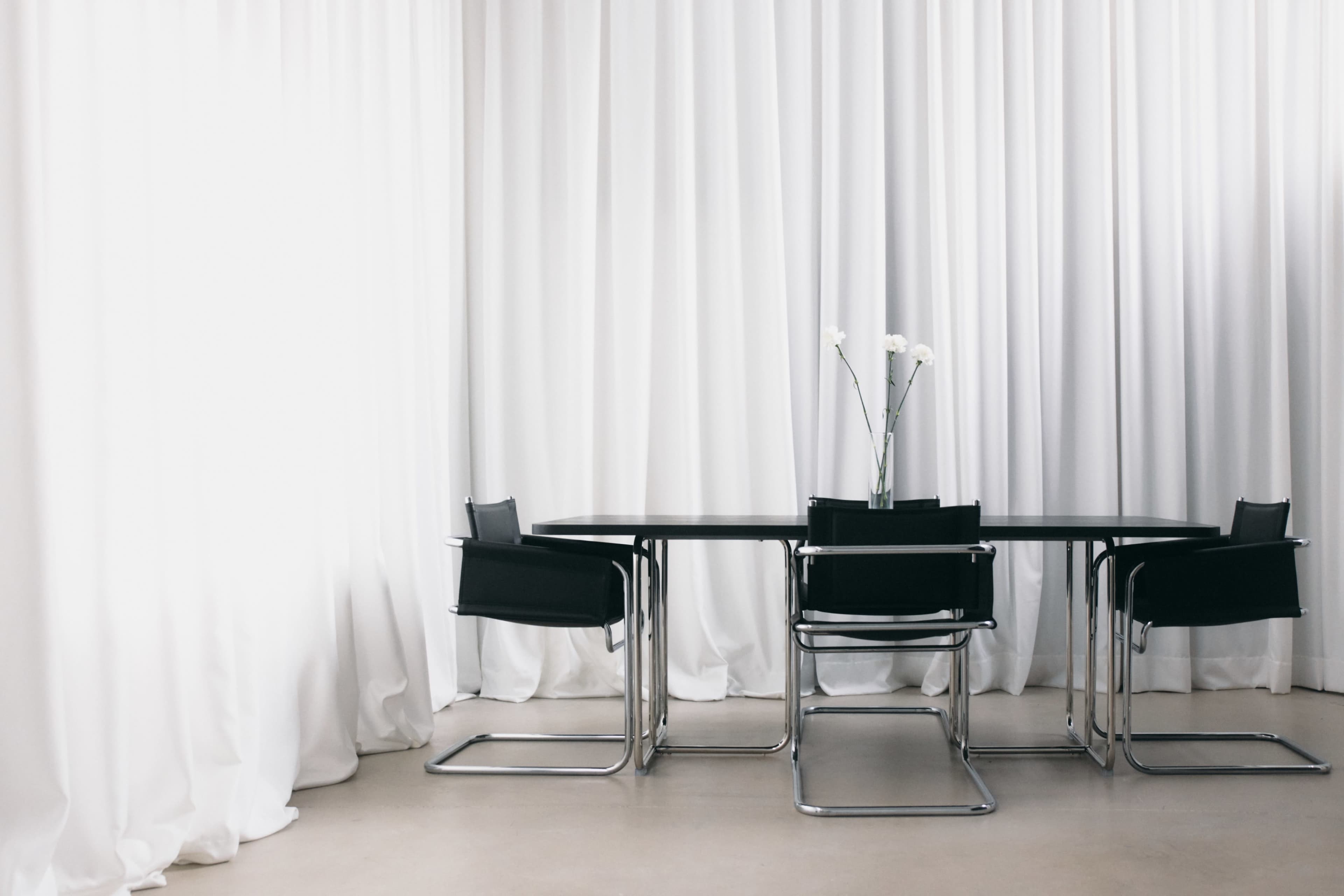 A rectangular black table with four black chairs surrounded by white curtains and a vase of flowers on the table.
