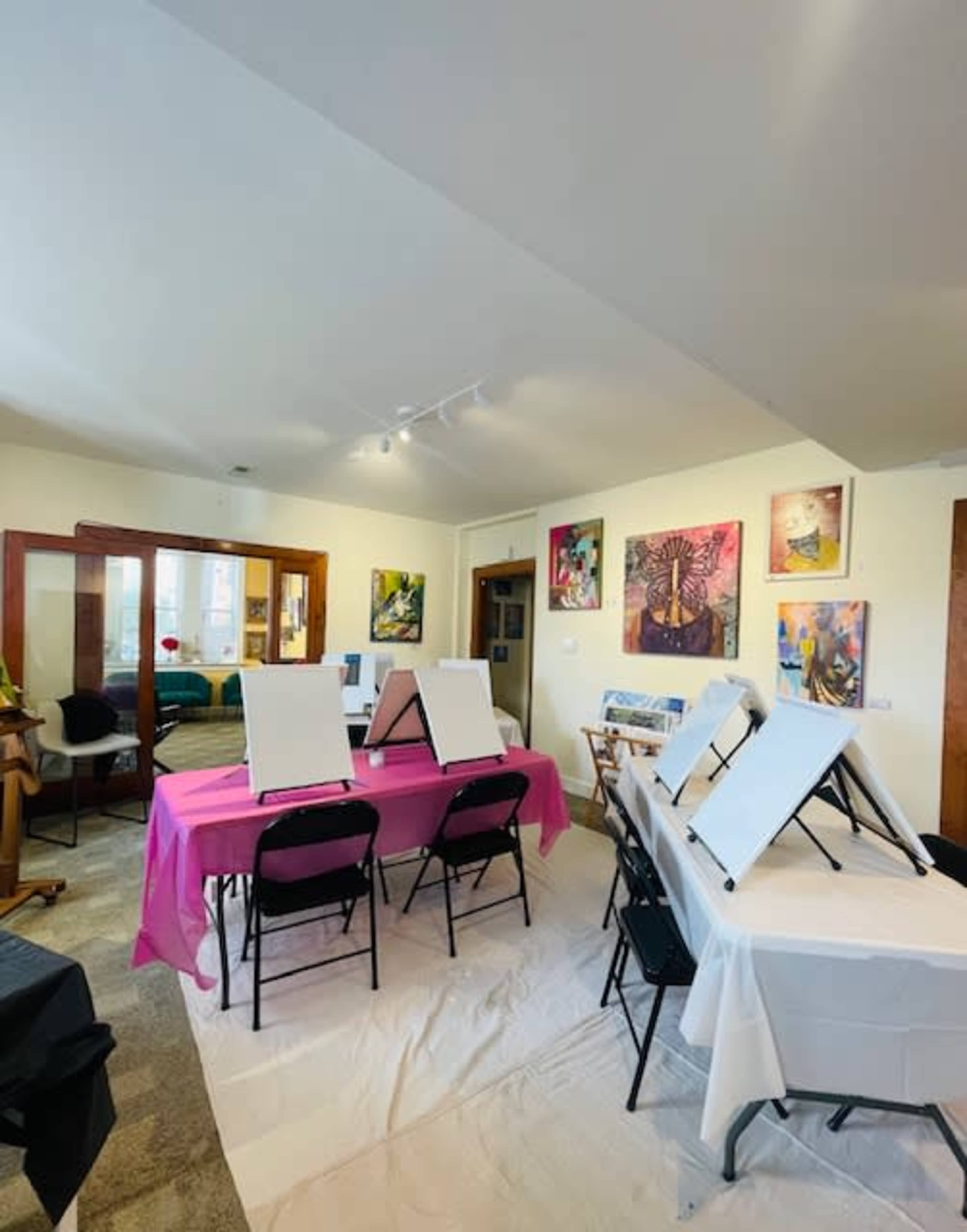 The image shows an art studio setup with tables covered in pink and white cloths, easels, and various artworks displayed on the walls.
