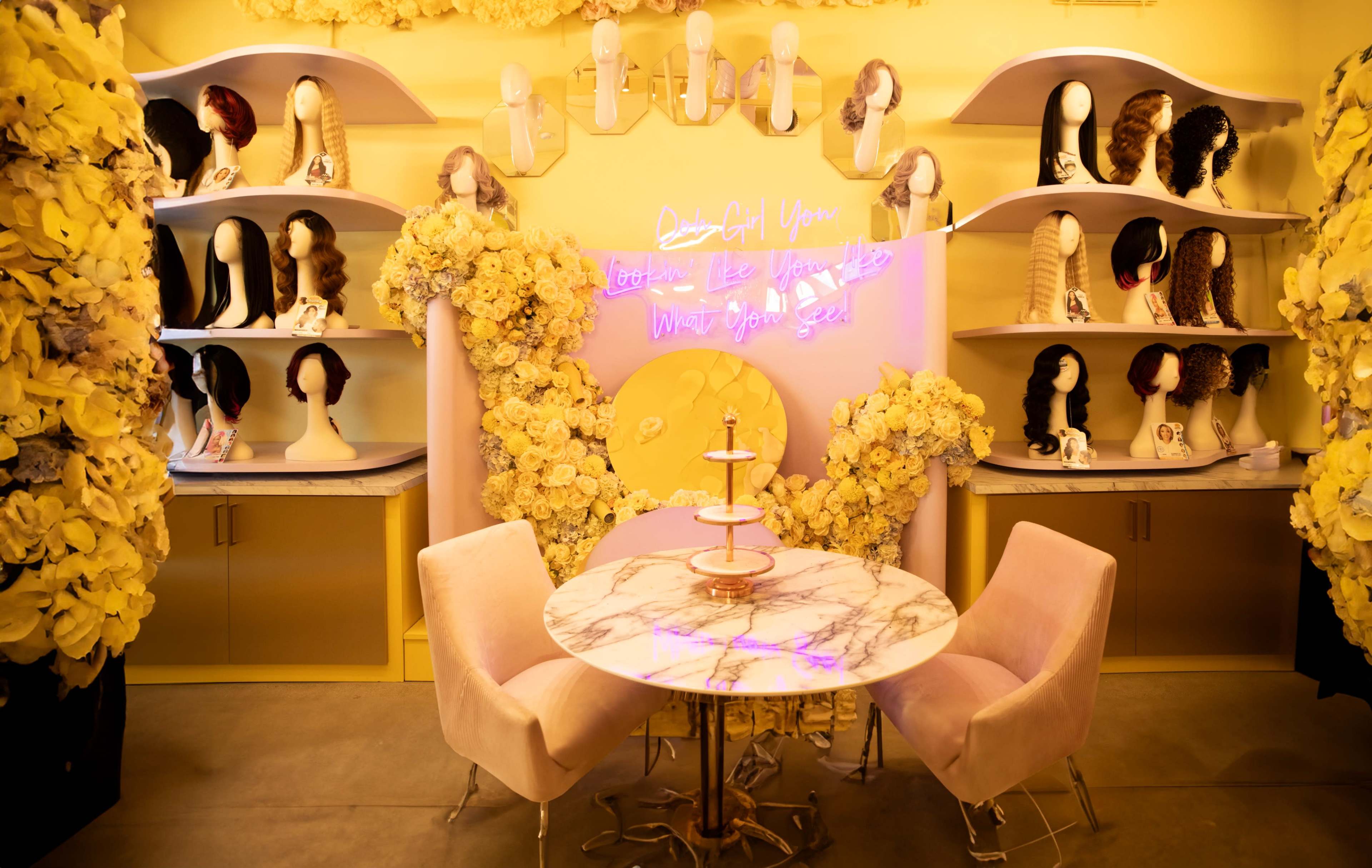 The image shows a brightly lit salon space adorned with yellow flowers and shelves displaying various wigs, accompanied by a round marble table and pink chairs.