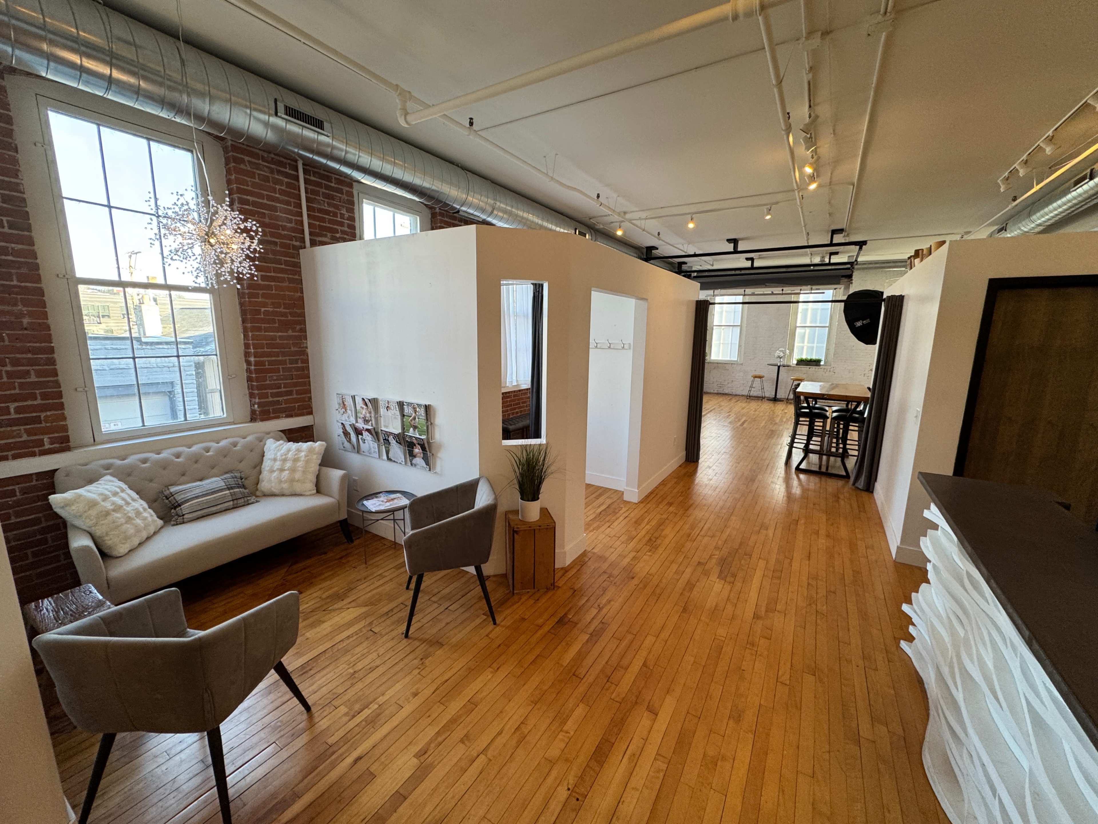 The image shows a modern office space with wooden flooring, a cozy seating area, partitioned workspaces, and large windows allowing natural light.