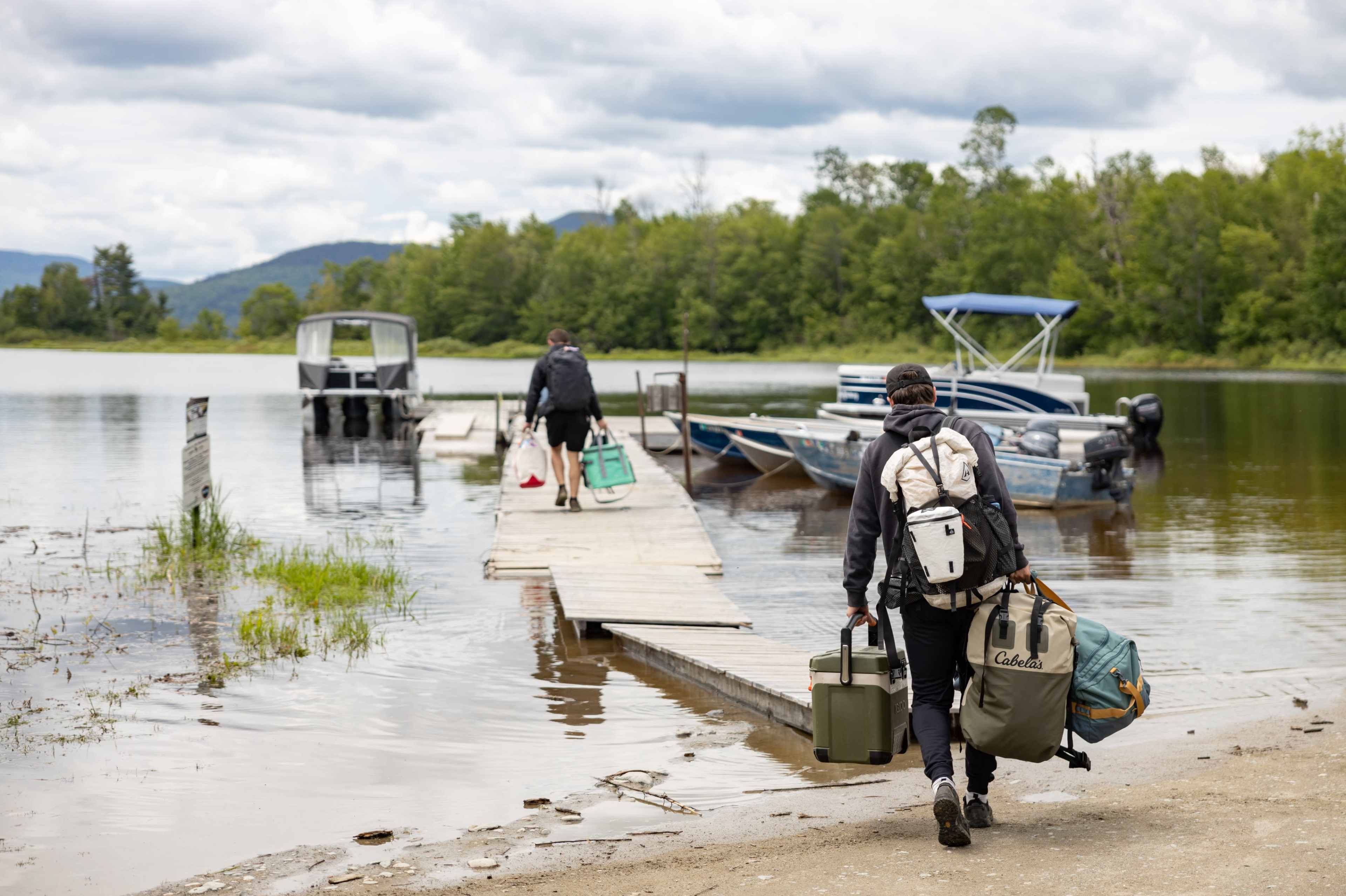 Two individuals carry gear along a dock toward a group of boats on a calm lake surrounded by trees and mountains.