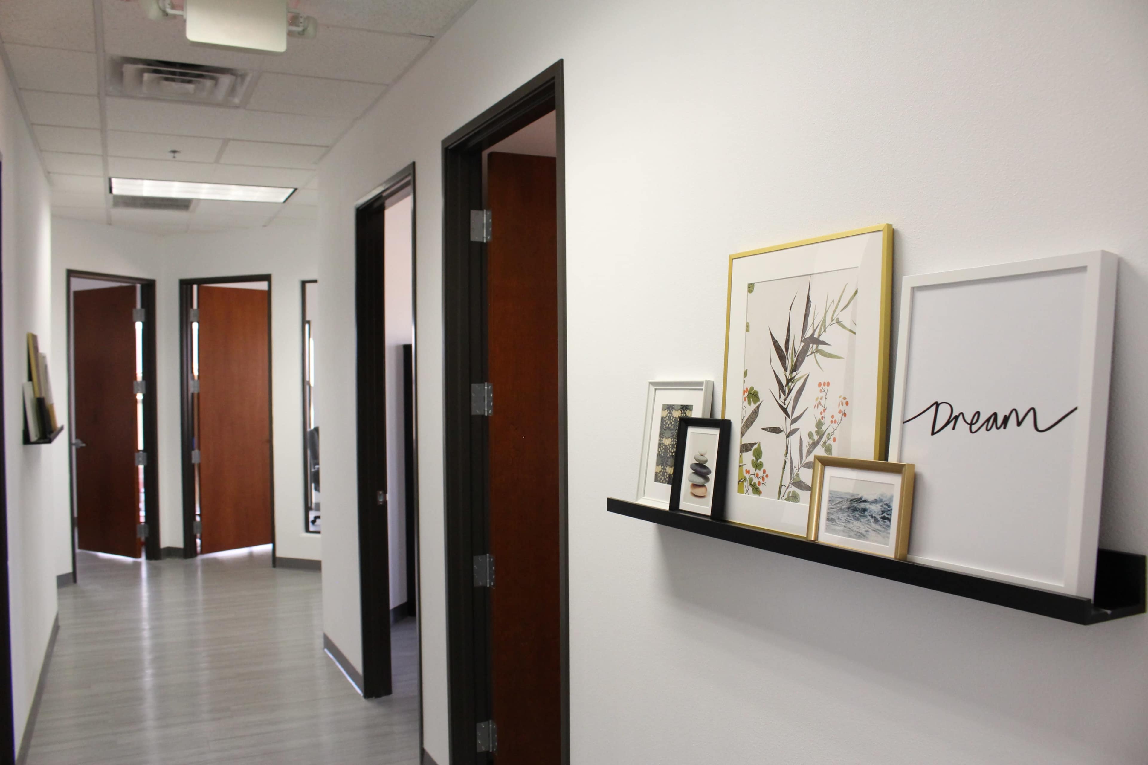 A modern office hallway featuring framed artwork and a shelf with decorative items along the wall, with open doors leading to other office spaces.