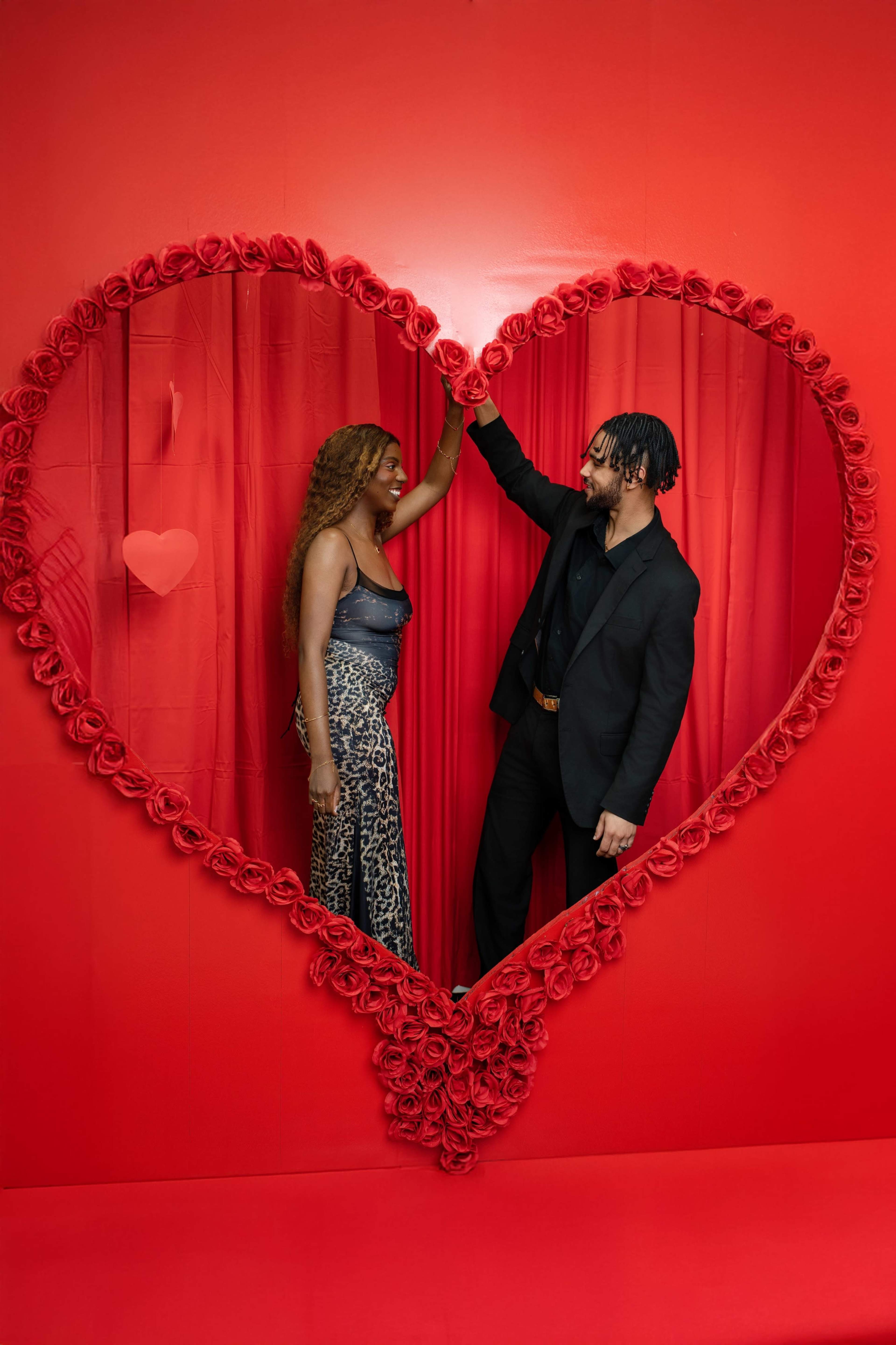 A couple poses playfully inside a large heart-shaped frame adorned with roses against a red backdrop.