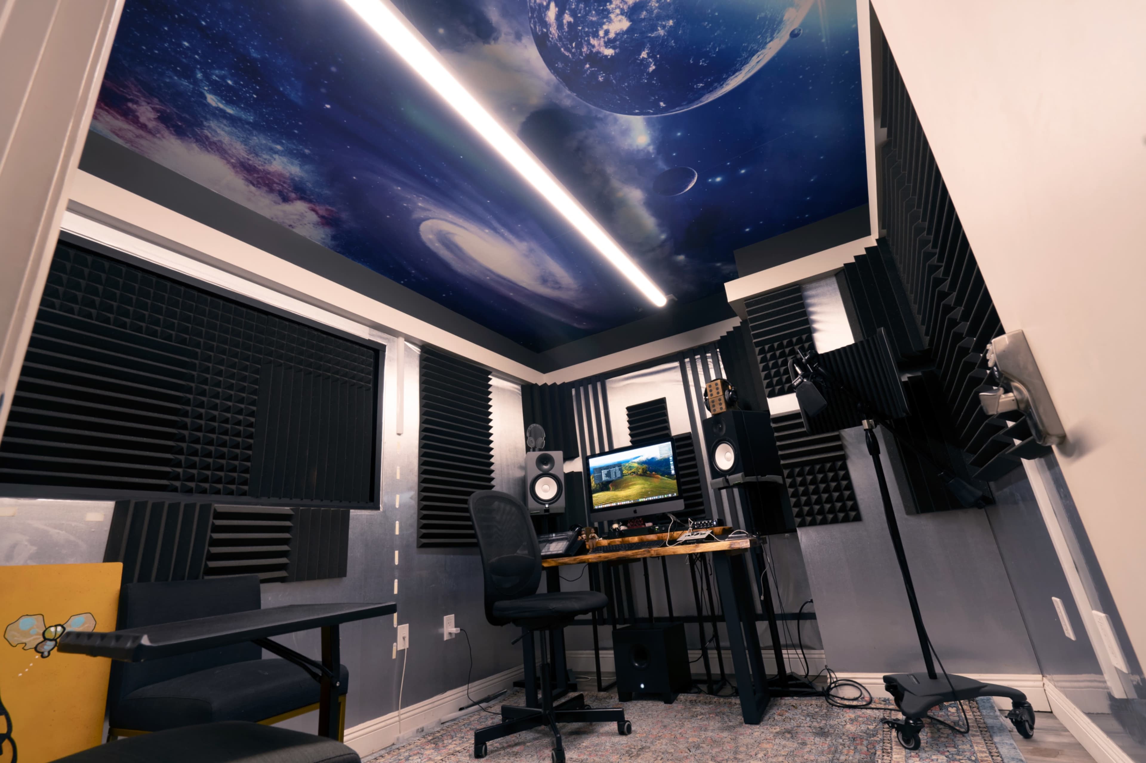 The image shows a recording studio equipped with soundproofing panels, a desk with audio equipment, and a ceiling featuring a cosmic mural.