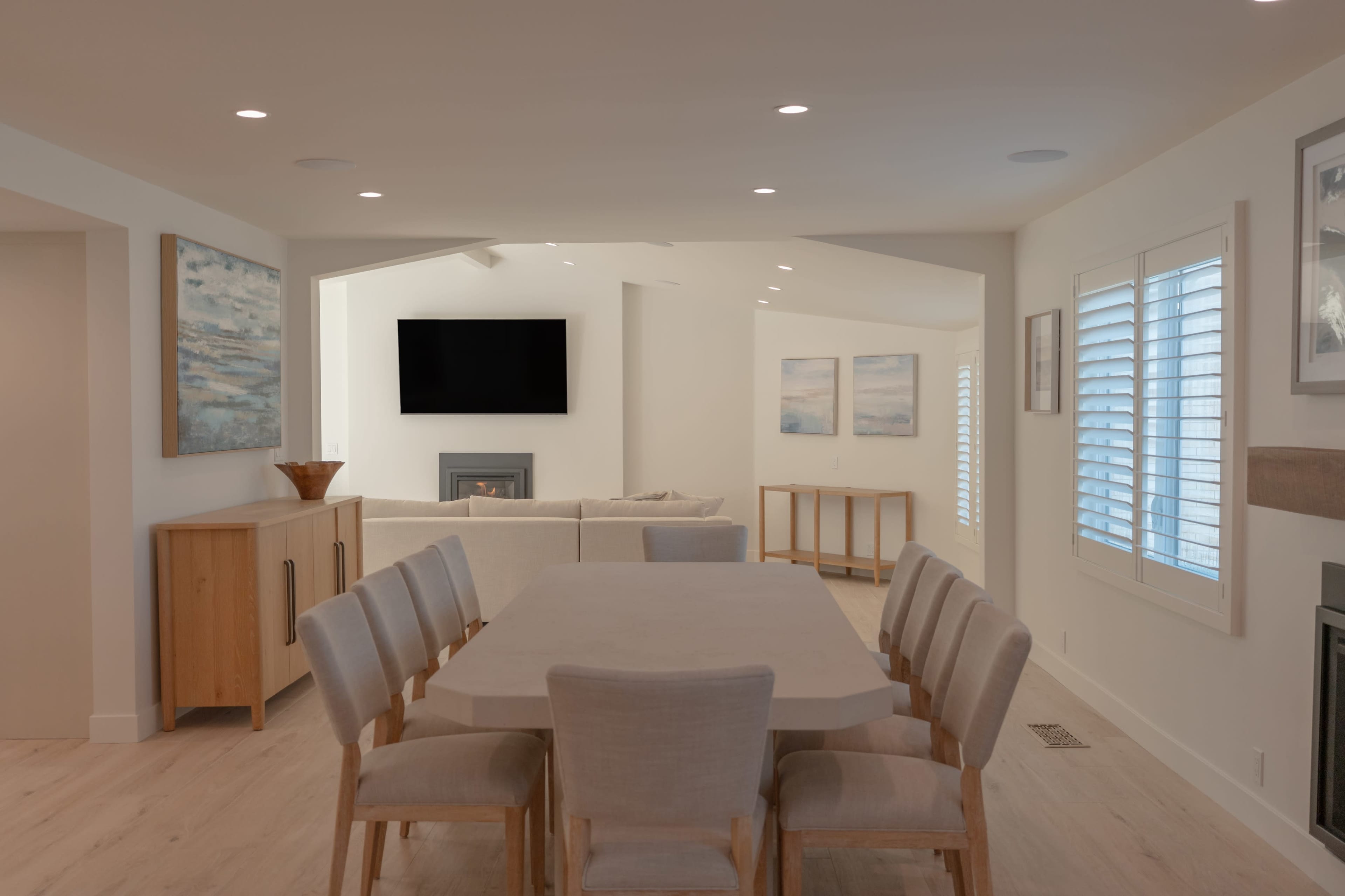 A dining area with a large table and chairs is connected to a bright, open living space featuring a television and artwork on the walls.