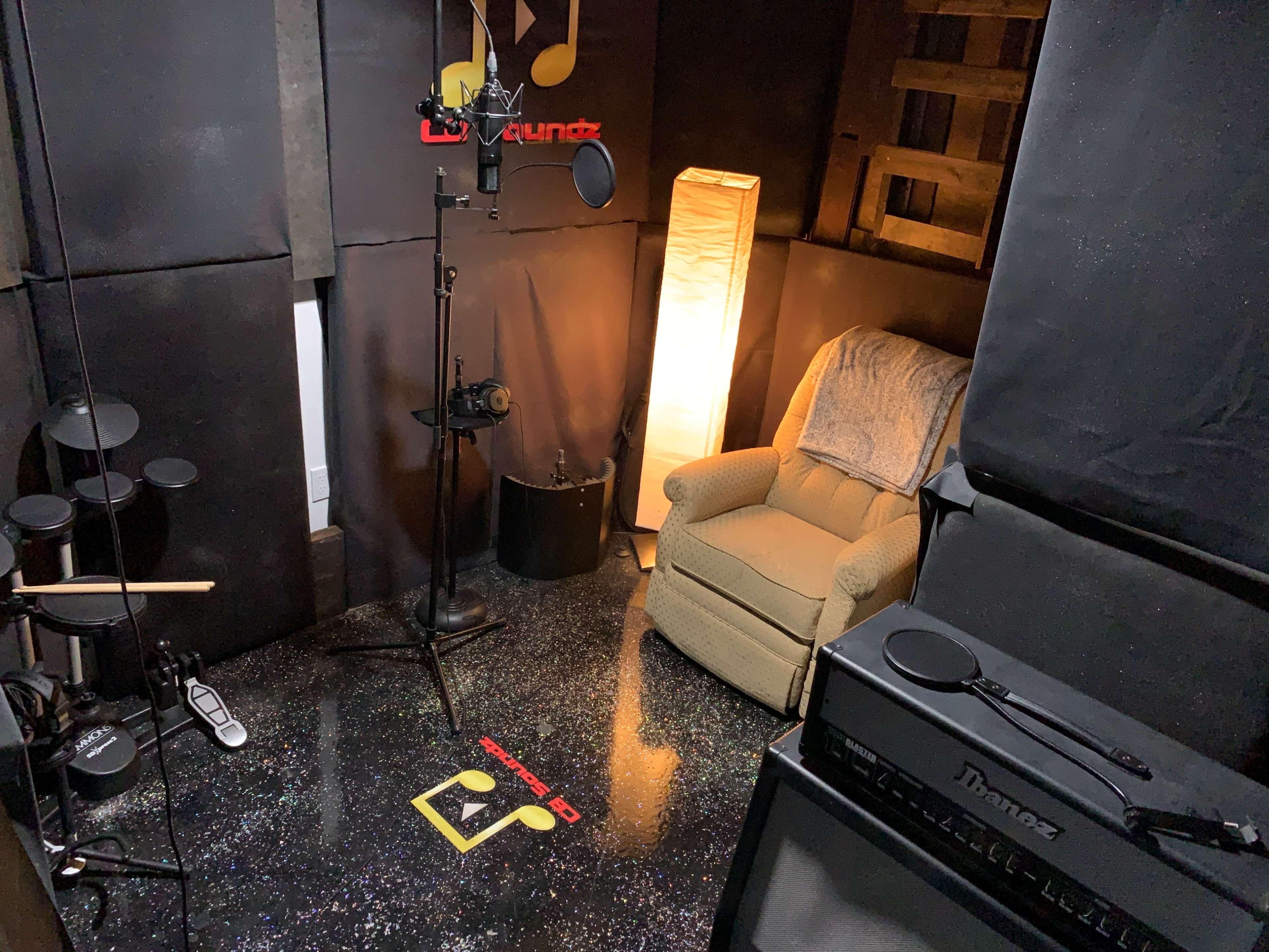 The image shows a small recording studio with a microphone on a stand, a chair, and a floor illuminated by a lamp.