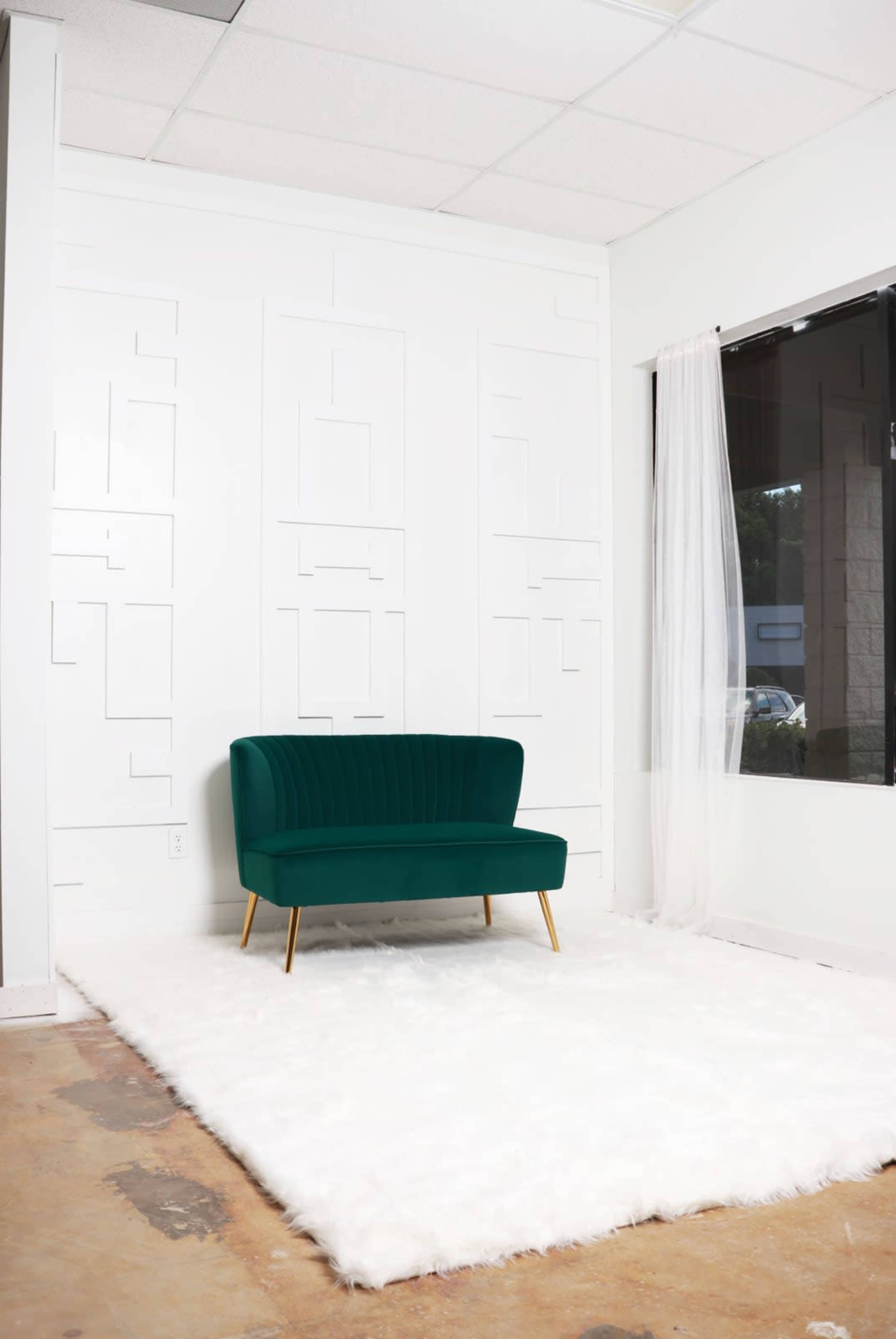 A green velvet loveseat is positioned on a white, fluffy rug in a minimally decorated room with a white wall and a large window.