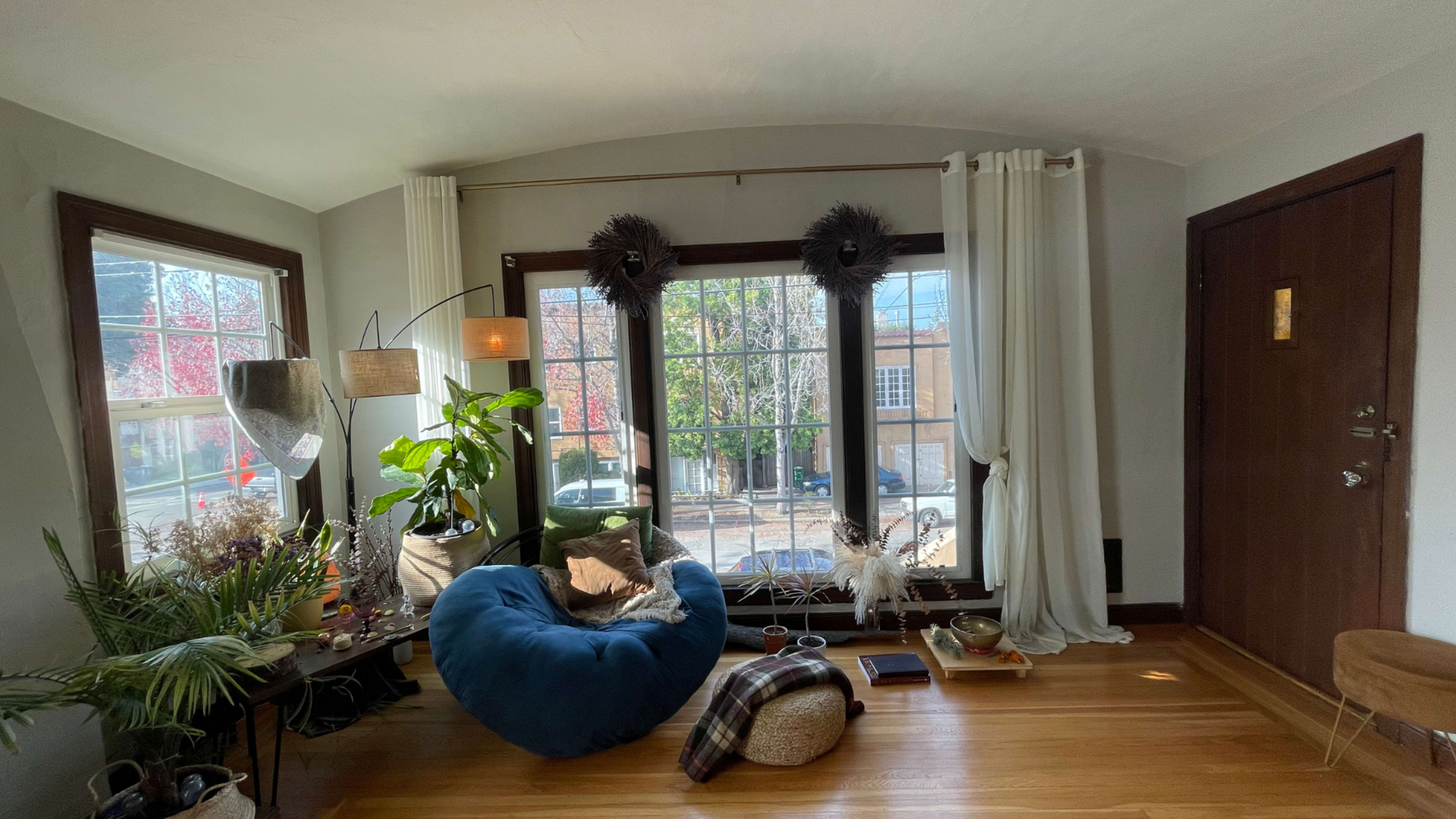 A cozy living room features a blue bean bag chair, potted plants, and large windows letting in natural light.