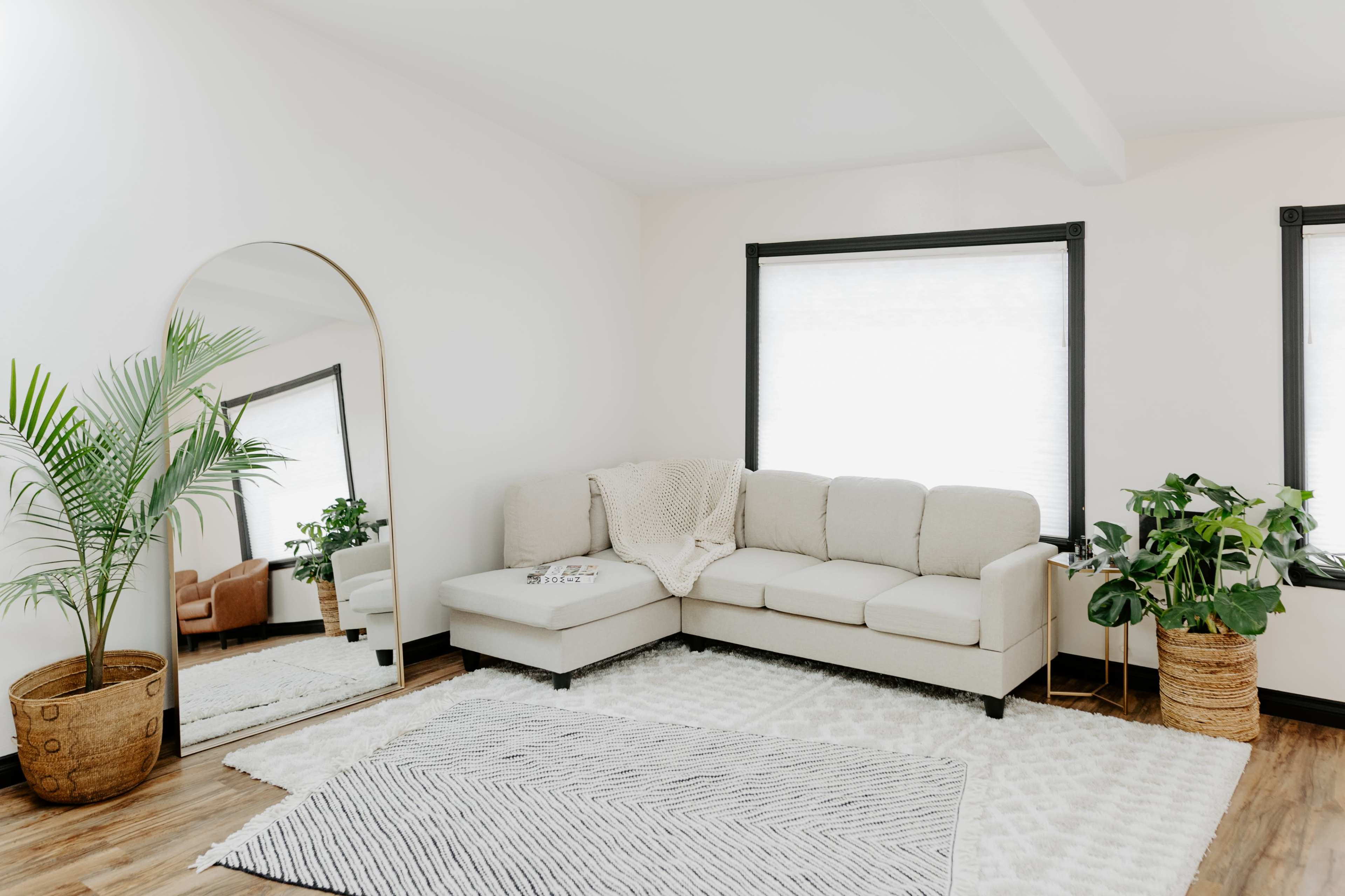 The scene features a modern living room with a beige sectional sofa, a large mirror, and potted plants beside two windows.