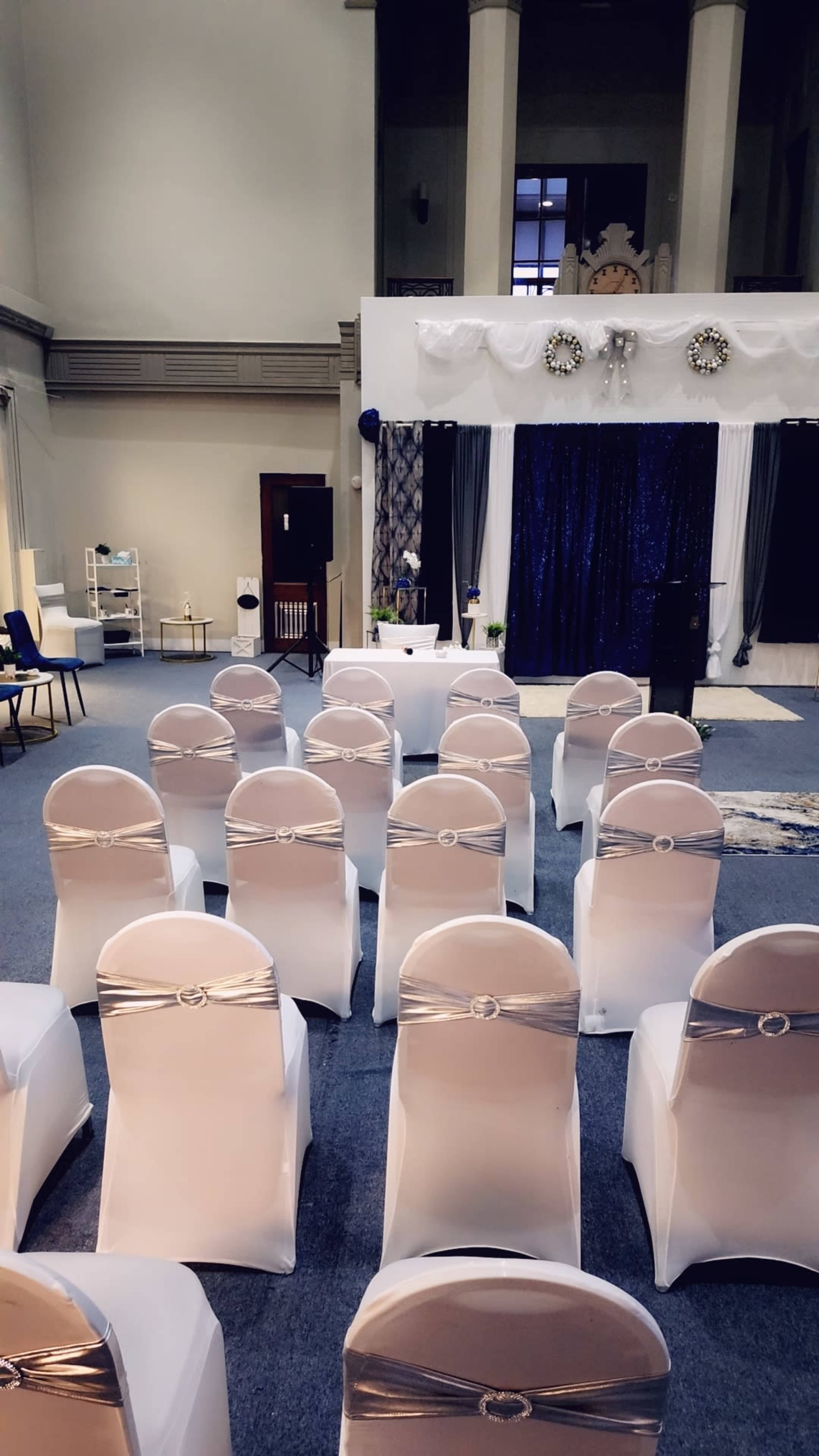 A hall set up for an event, with rows of white chairs adorned with silver sashes facing a decorated stage.