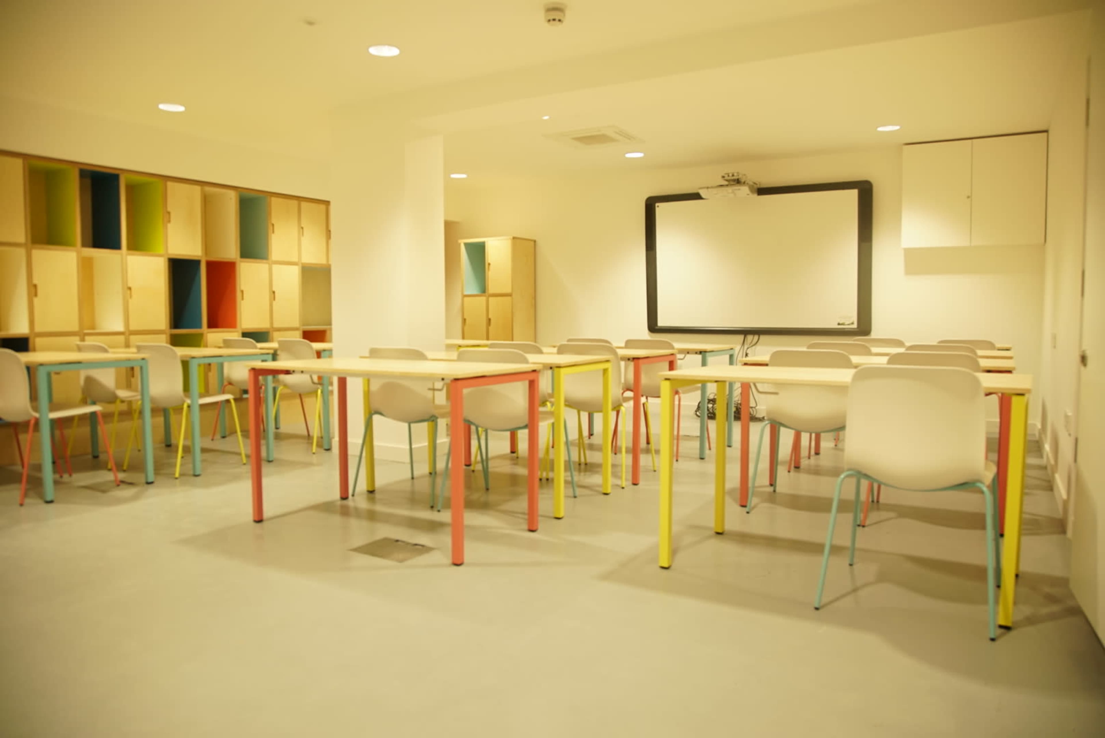 A brightly colored classroom with multiple tables and chairs arranged around a projector screen and storage cabinets.