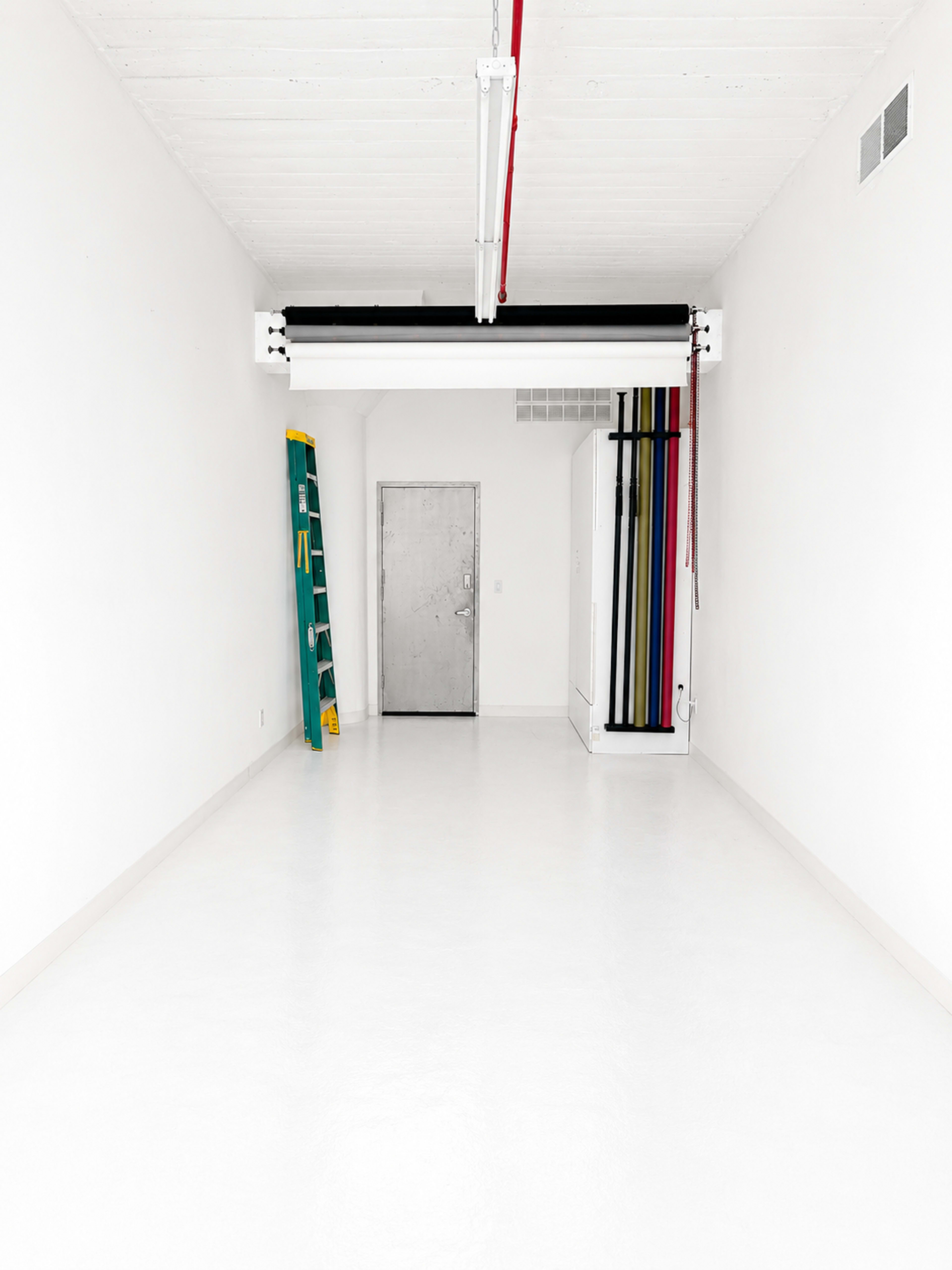 The image shows a spacious, empty room with white walls and a polished white floor, featuring a metal door, a ladder against one wall, and a series of colored lines or cords hanging from the ceiling.