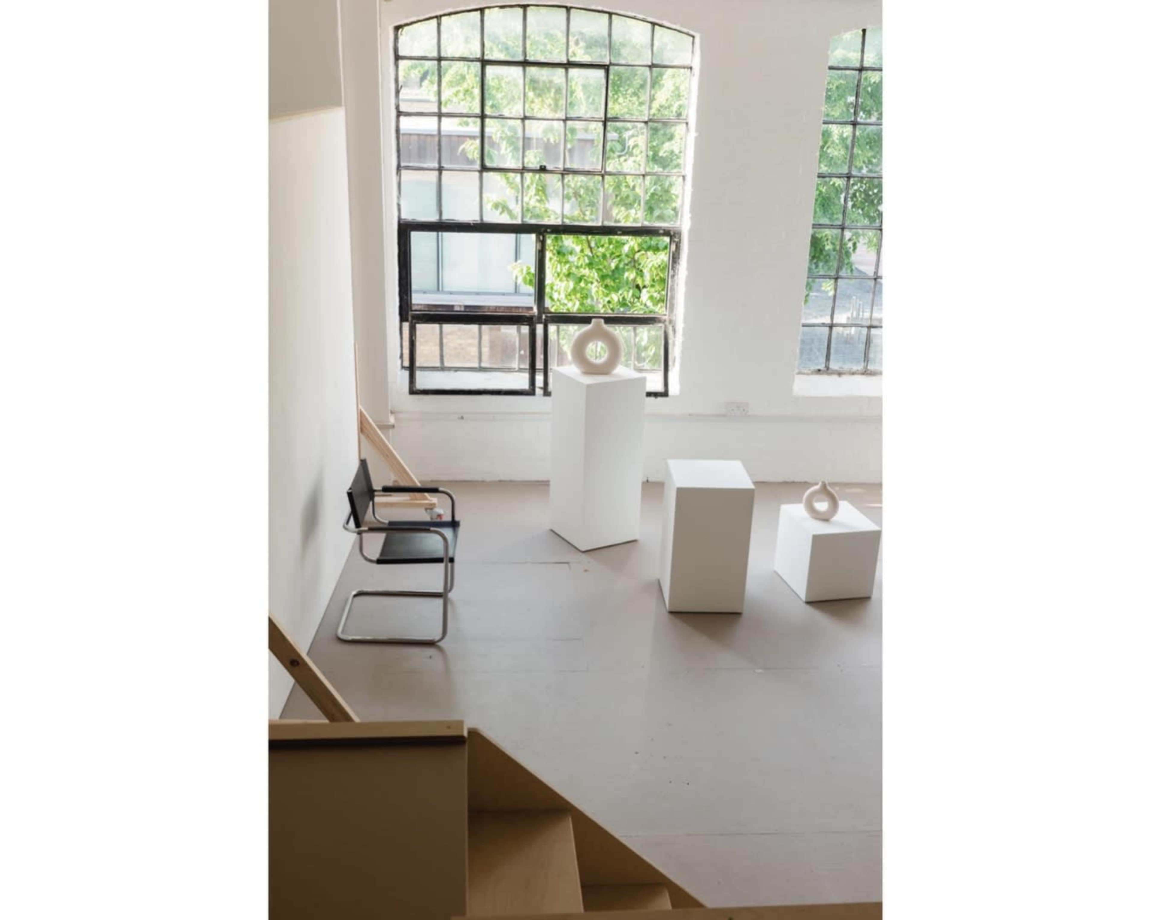 A minimalistic art gallery with large windows, featuring white sculptures on pedestals and a black chair in the foreground.