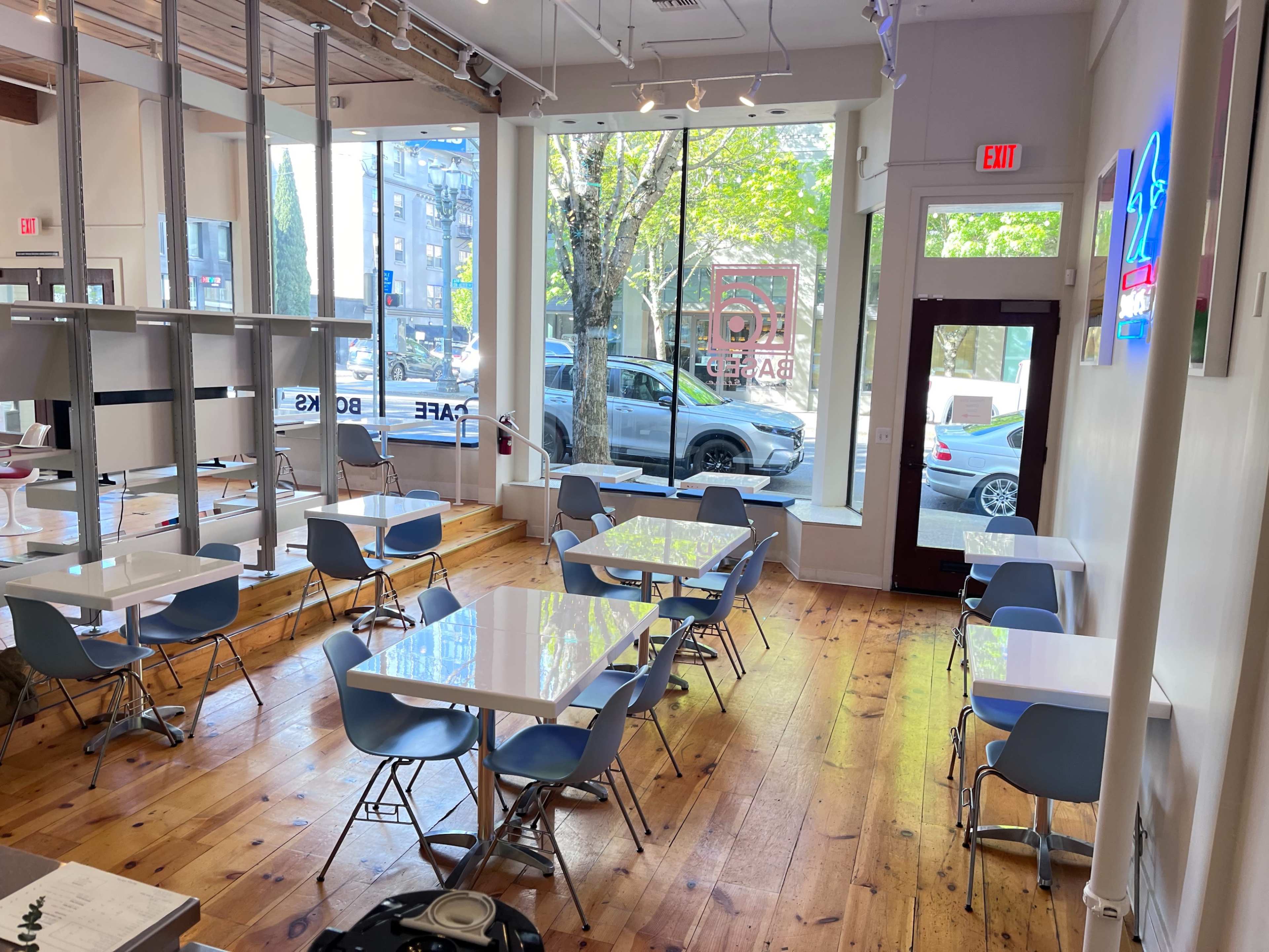 The image shows a modern café with white tables and blue chairs, featuring large windows that let in natural light and a view of trees and parked cars outside.