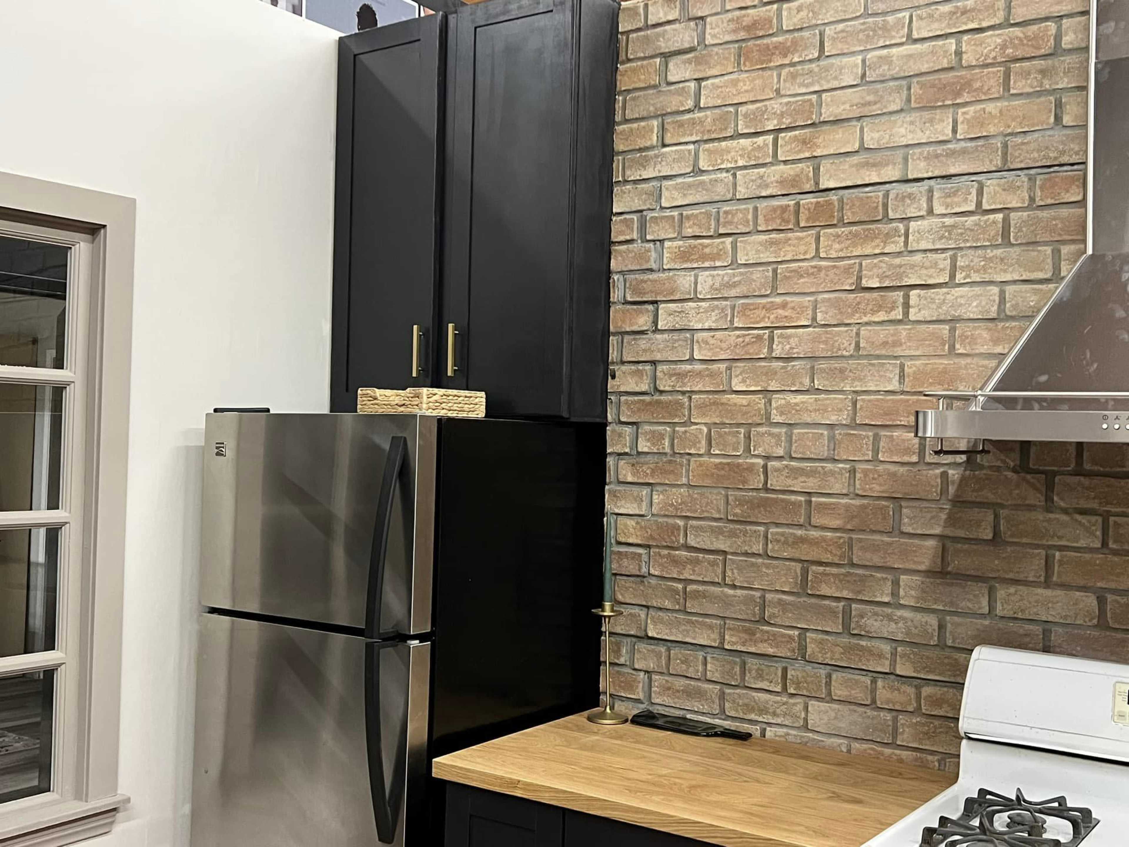 A kitchen corner with a stainless steel refrigerator, a wooden countertop, and a brick wall covered by dark cabinetry.
