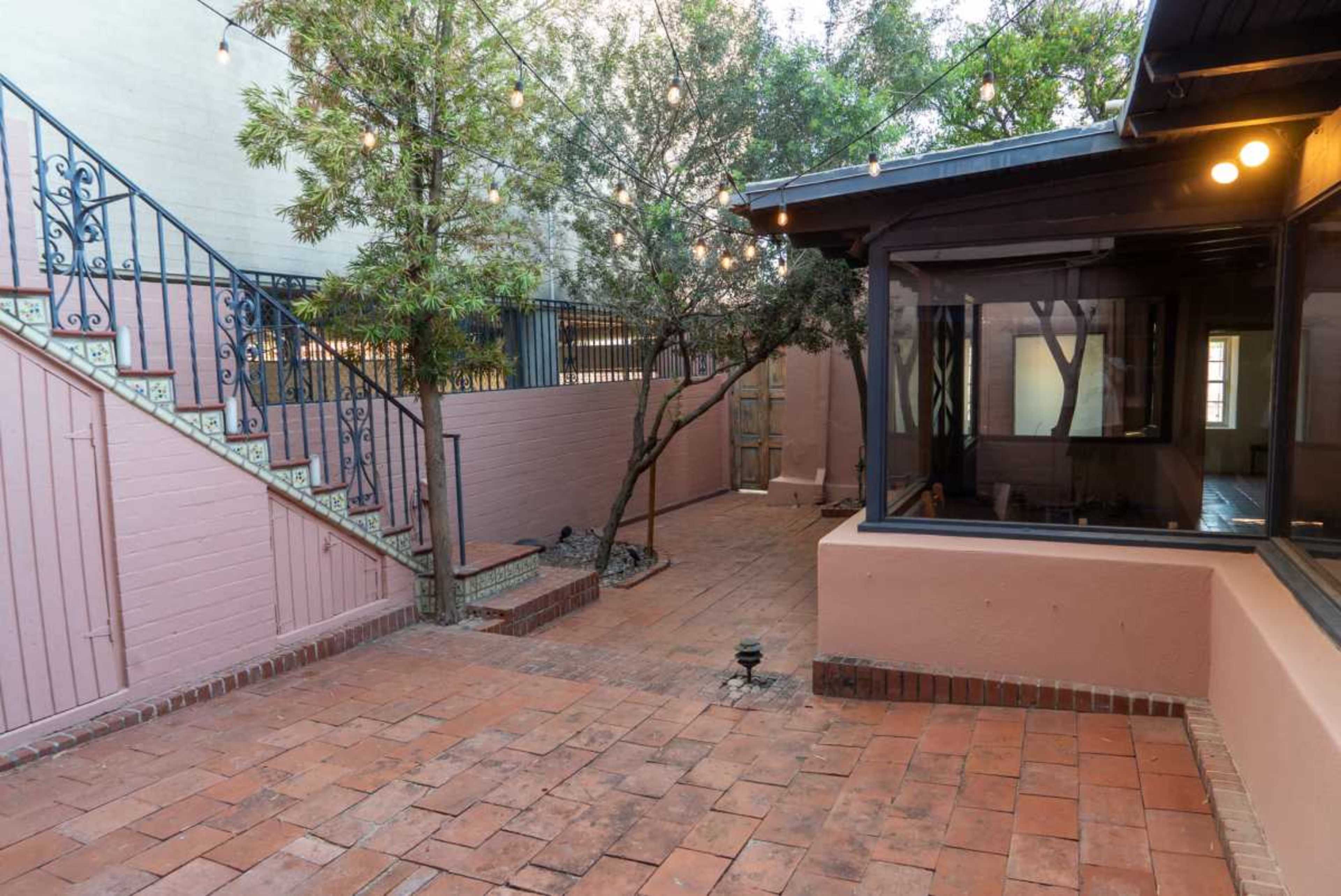 The image shows a courtyard with brick paving, surrounded by walls, a stairway with decorative railings, and a small building with windows.