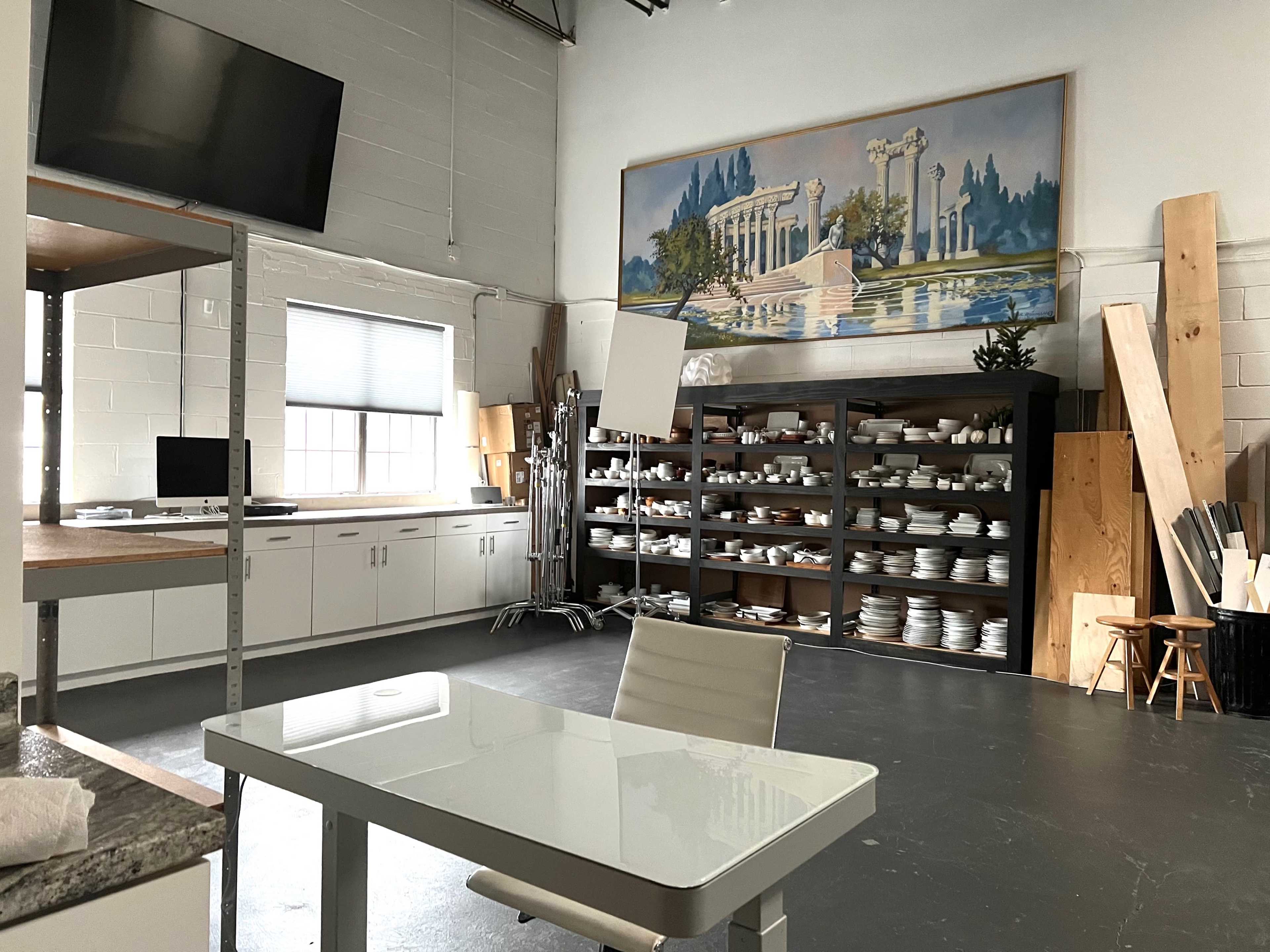 A spacious studio with a white table and chair in front, shelves filled with dishes in the background, and a large mural depicting classical architecture on the wall.