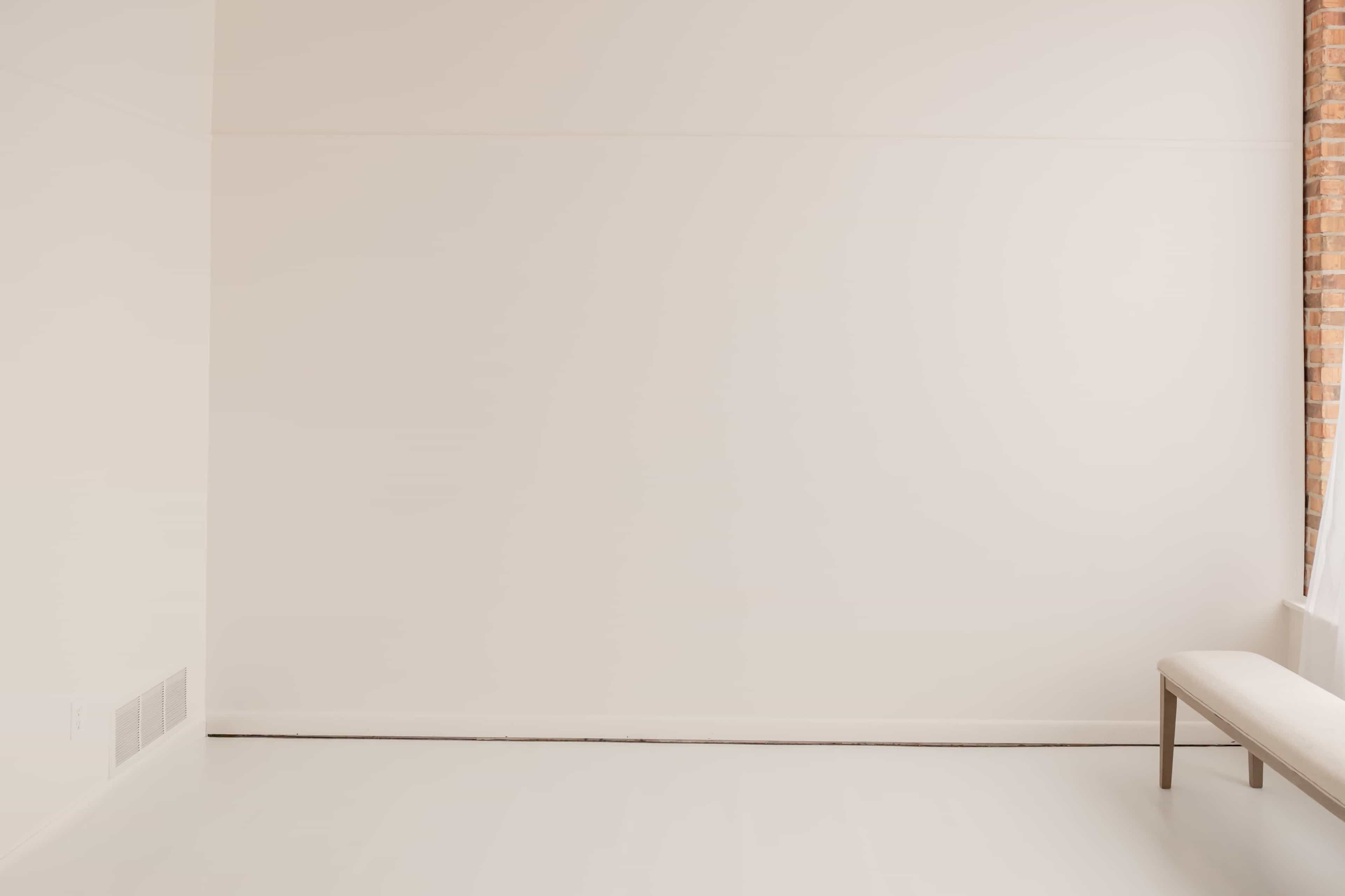 The image shows a minimalistic room with a blank white wall and a simple bench against the wall.